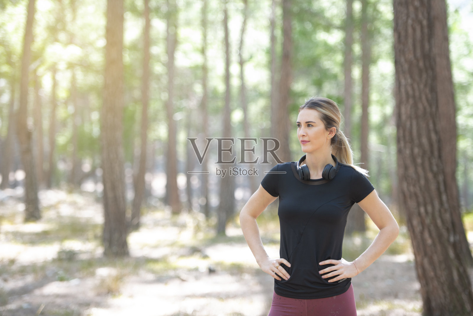 在森林里做运动的妇女正在训练后休息。金发女运动员戴着耳机，双手叉腰。照片摄影图片