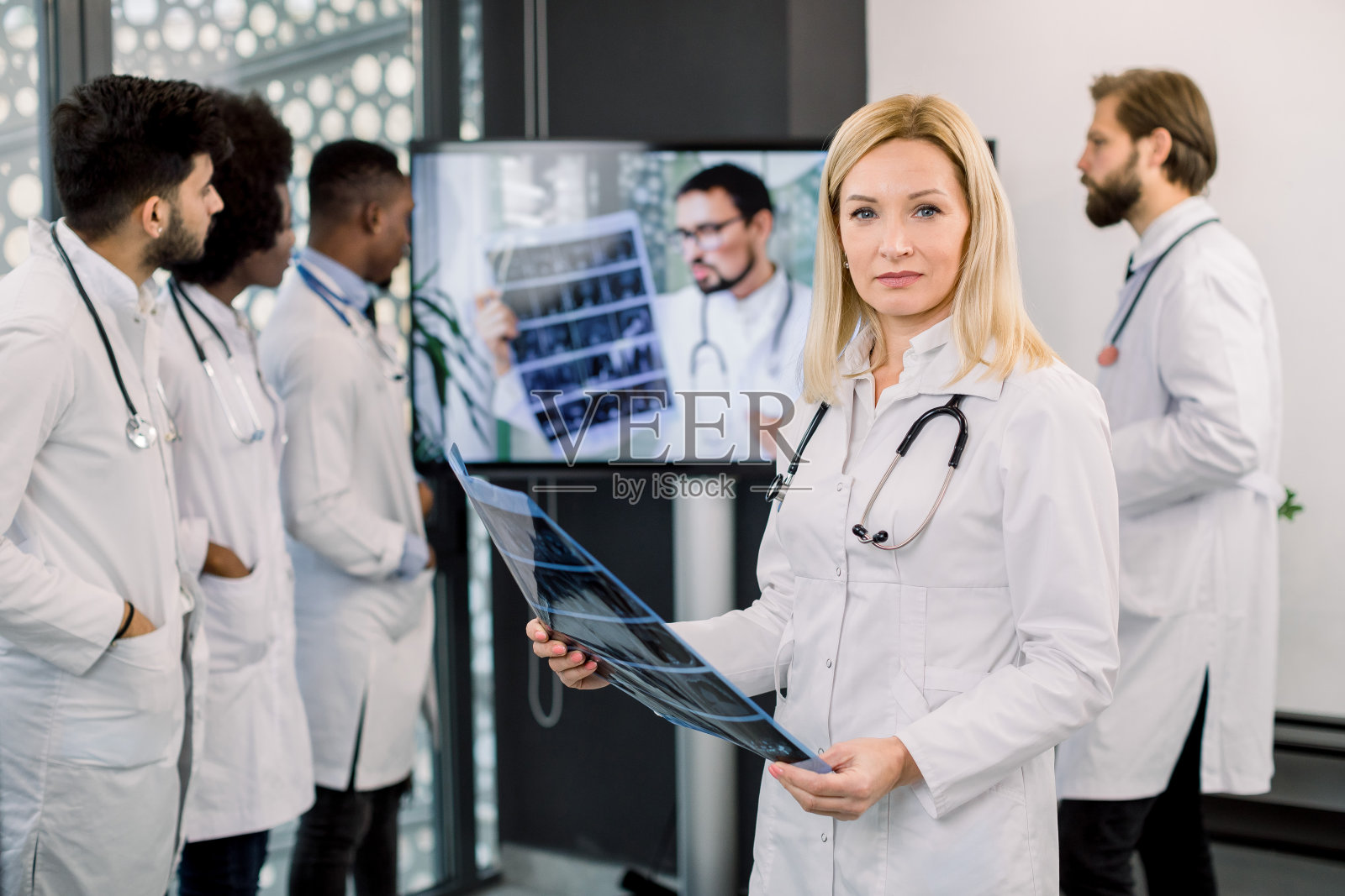 美丽成熟的金发女医生的肖像拿着CT扫描，看着相机。她的多种族同事们在大数字屏幕上听男性专家讨论计算机断层扫描的结果照片摄影图片