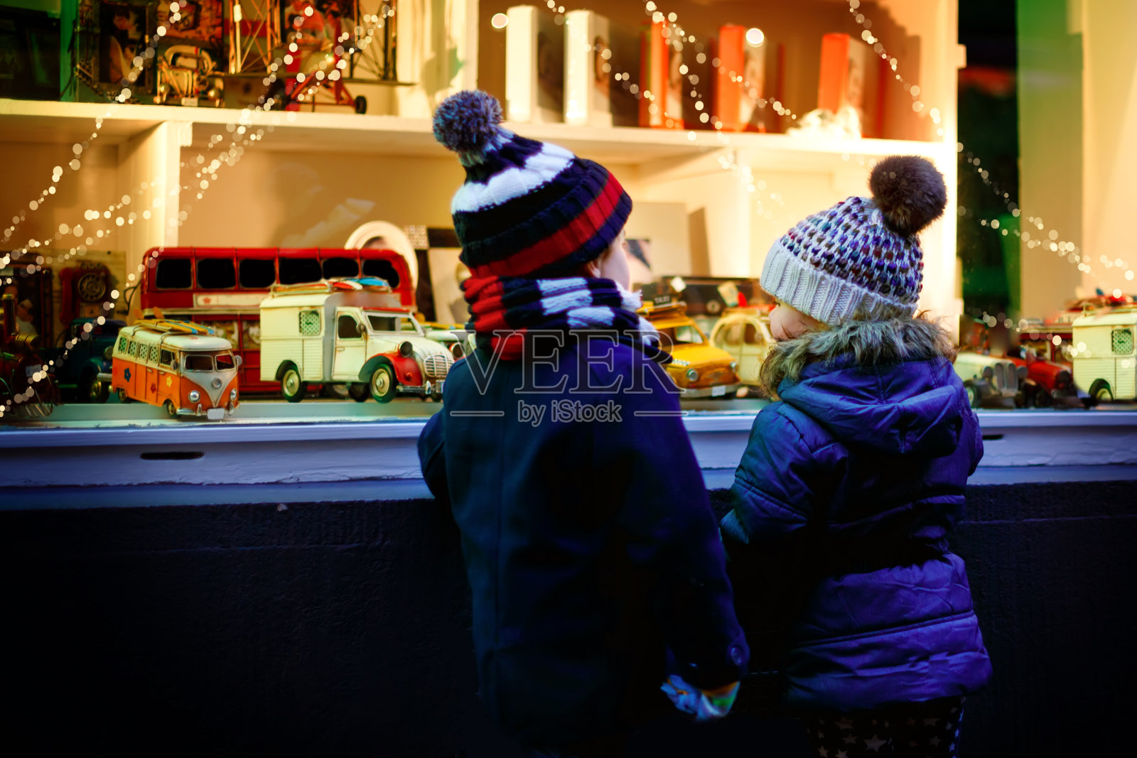 两个刚学走路的孩子，一男一女在圣诞节期间看橱窗里的汽车玩具。着迷的孩子们做梦和许愿。装饰着圣诞礼物的窗户。雪落下来。照片摄影图片