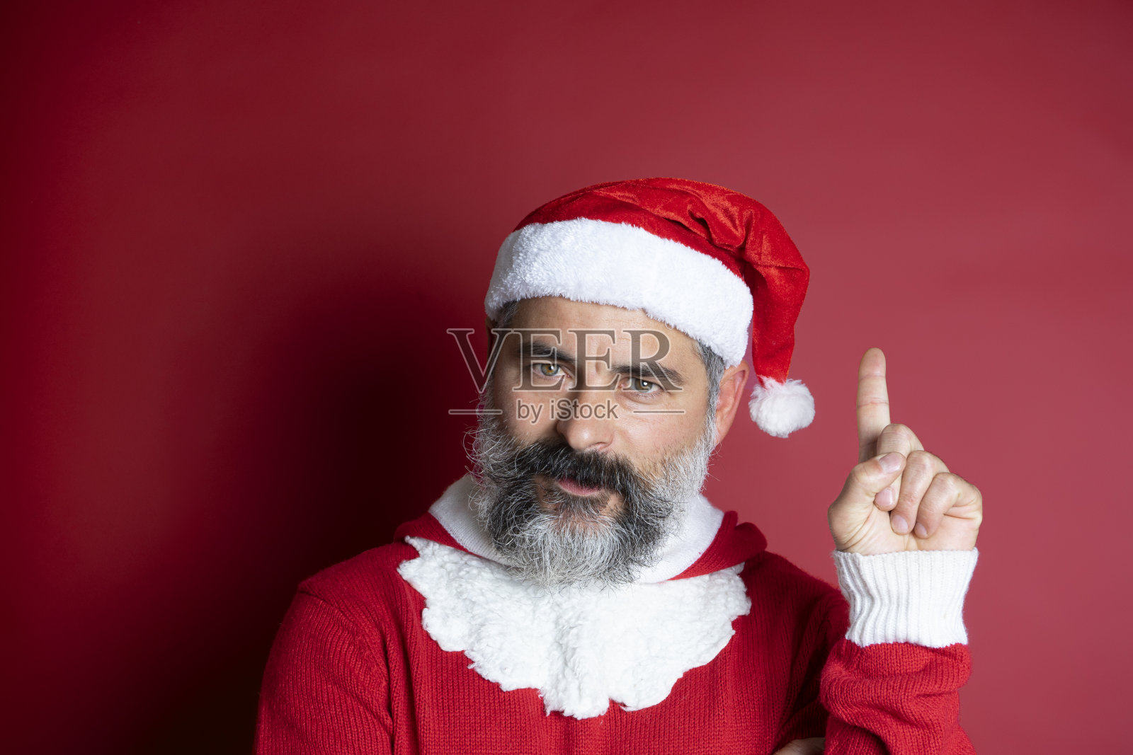 一个戴着圣诞帽的大胡子男人的特写，手指向上看着红色背景的摄像机照片摄影图片