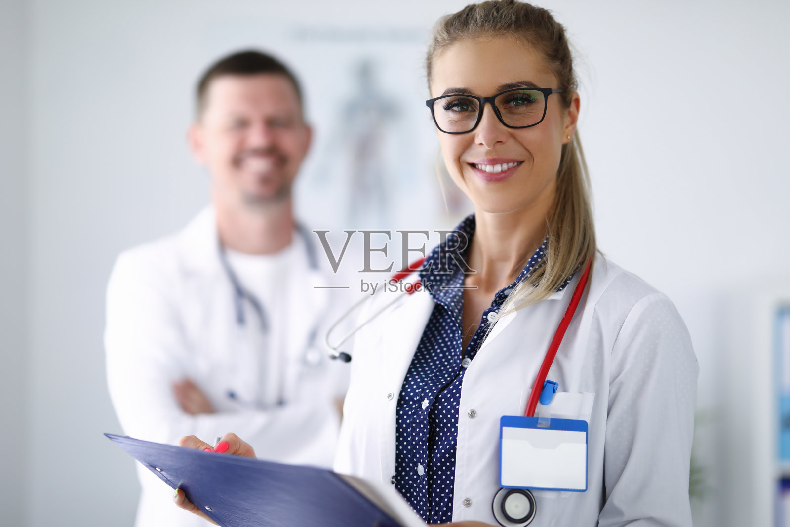 女医生微笑着拿着写字板站在她的同事身后。照片摄影图片