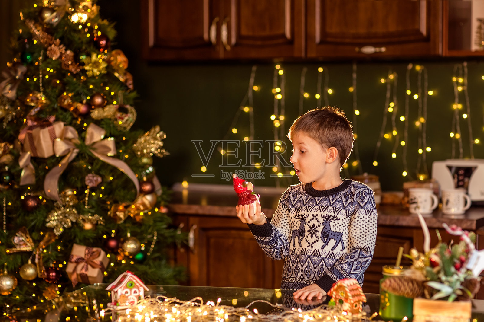 在装饰过的圣诞厨房里，一个男孩坐在桌子旁。拿着一个玩具装饰圣诞树。照片摄影图片