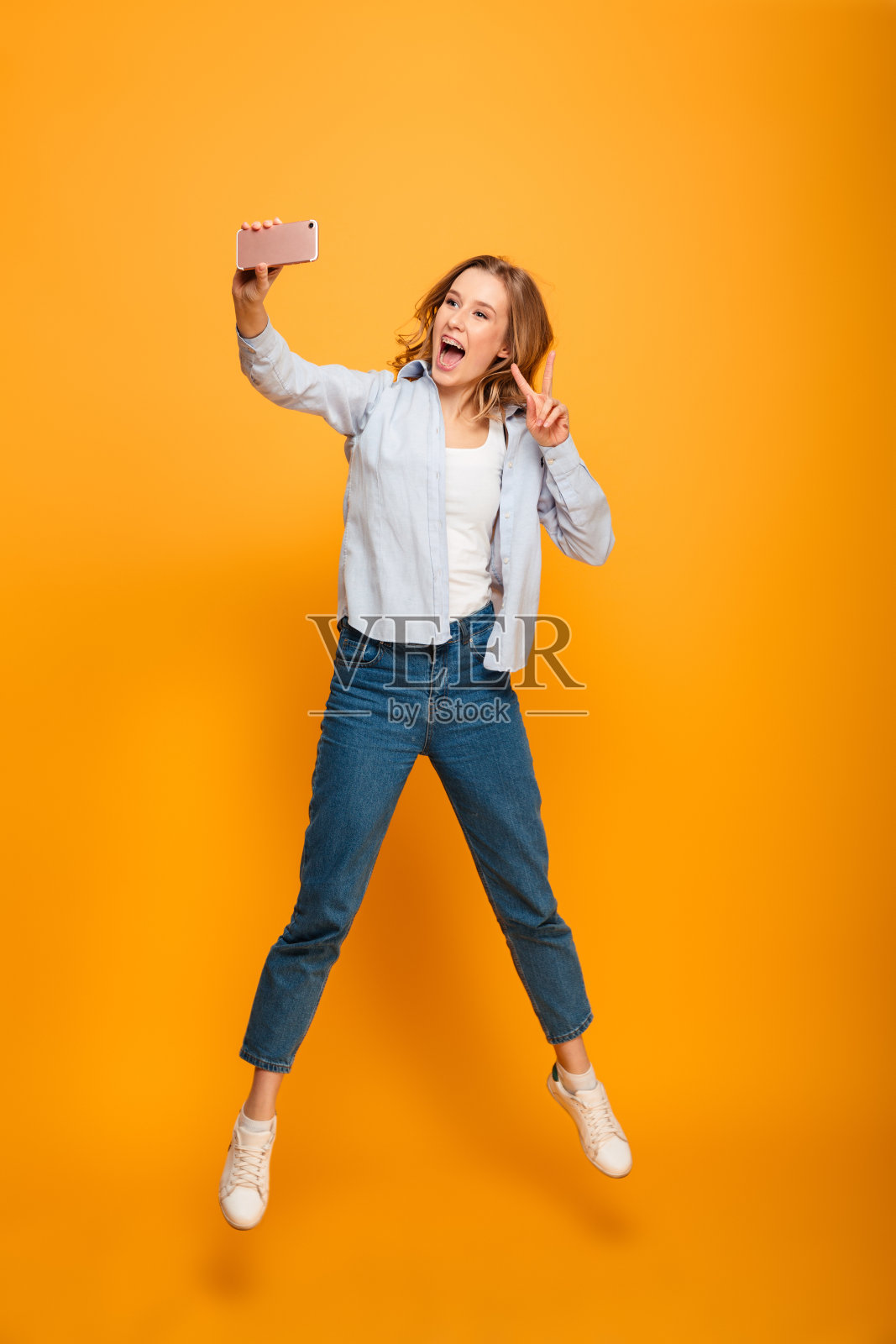 年轻迷人的女人的全长肖像微笑和显示胜利的标志，而用手机自拍，孤立在黄色的背景照片摄影图片