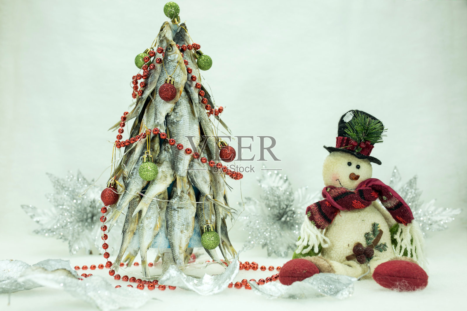 这棵圣诞树是用干鱼做的。祝渔夫新年快乐。一个快乐的雪人在树的旁边。背景装饰着圣诞节的银白色花朵。照片摄影图片