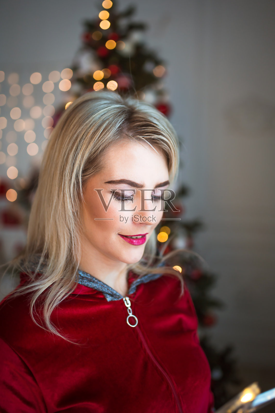 一个穿着红色运动服的金发女人站在圣诞树旁。在脸附近加花环。圣诞，新年，节日化妆，照片摄影图片