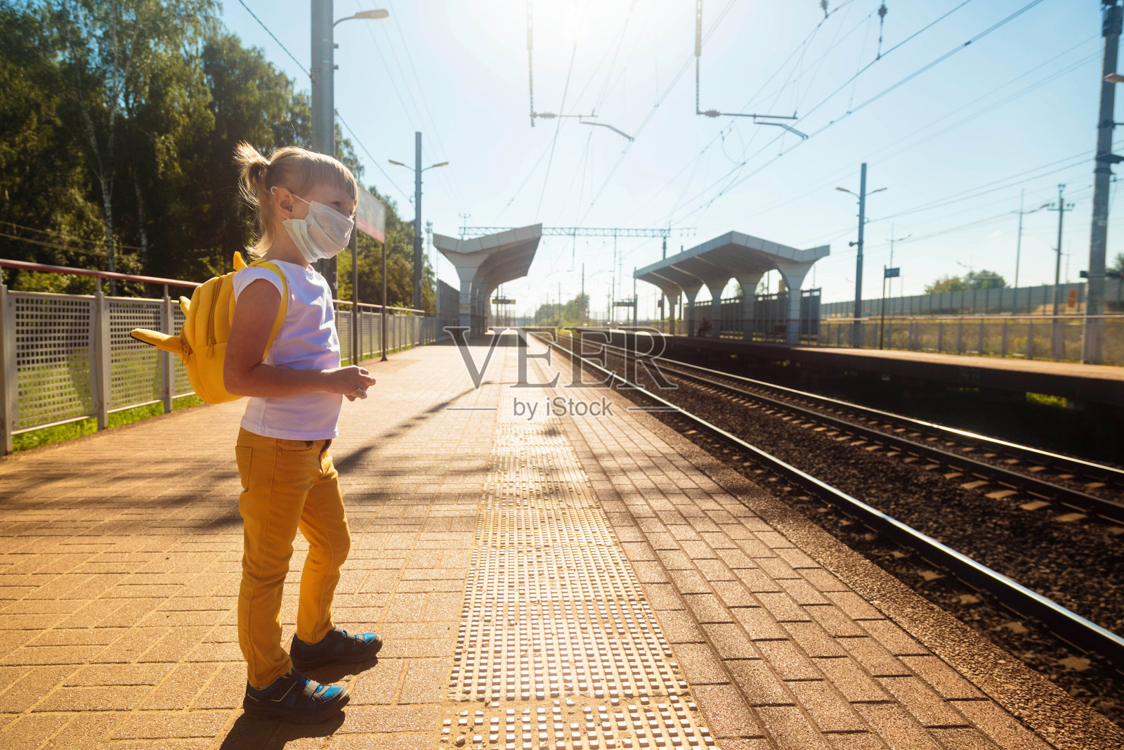 一个穿着白色t恤和牛仔裤带着医用口罩的小女孩在火车站等火车。保持社交距离，避免COVID-19传播。安全的旅行。照片摄影图片