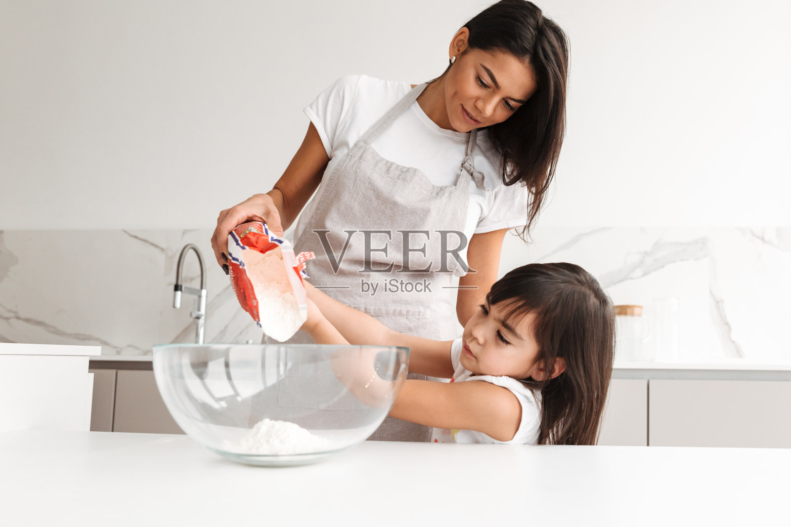 年轻的母亲穿着围裙和她的小女儿一起在家里的厨房里做糕点照片摄影图片