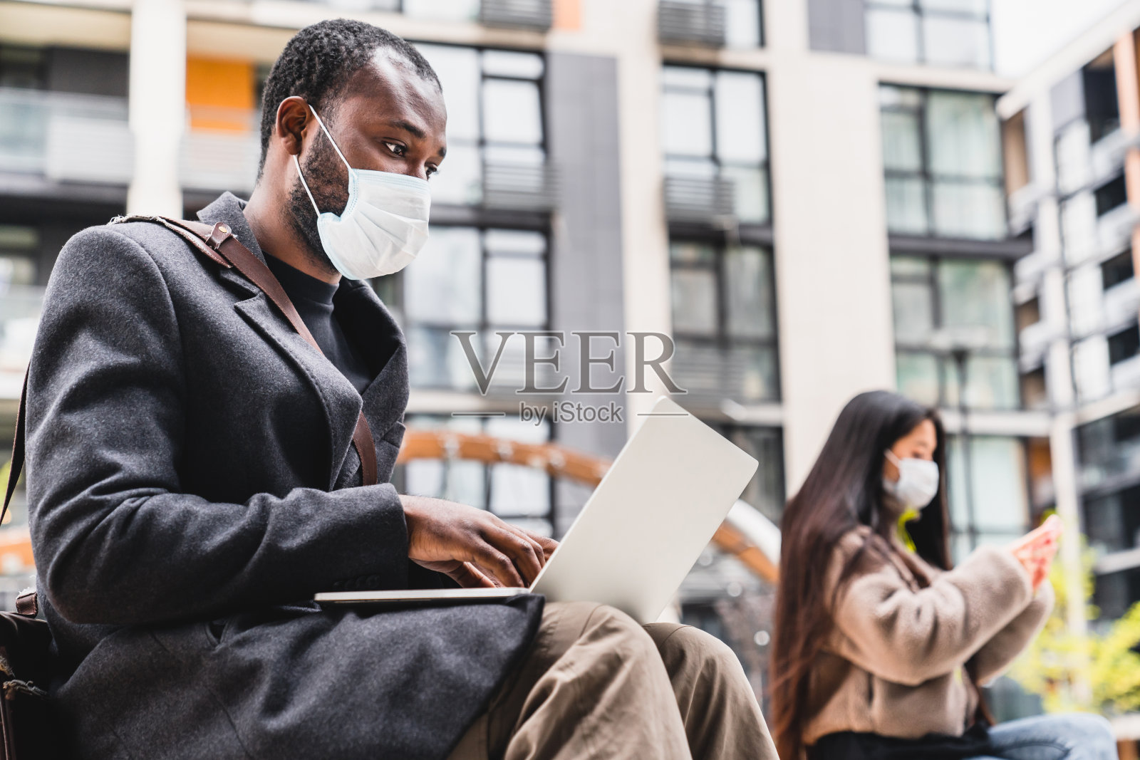 戴口罩的非洲男子使用笔记本电脑和戴口罩的亚洲女孩用手机保护自己免受2019冠状病毒感染的户外照片摄影图片