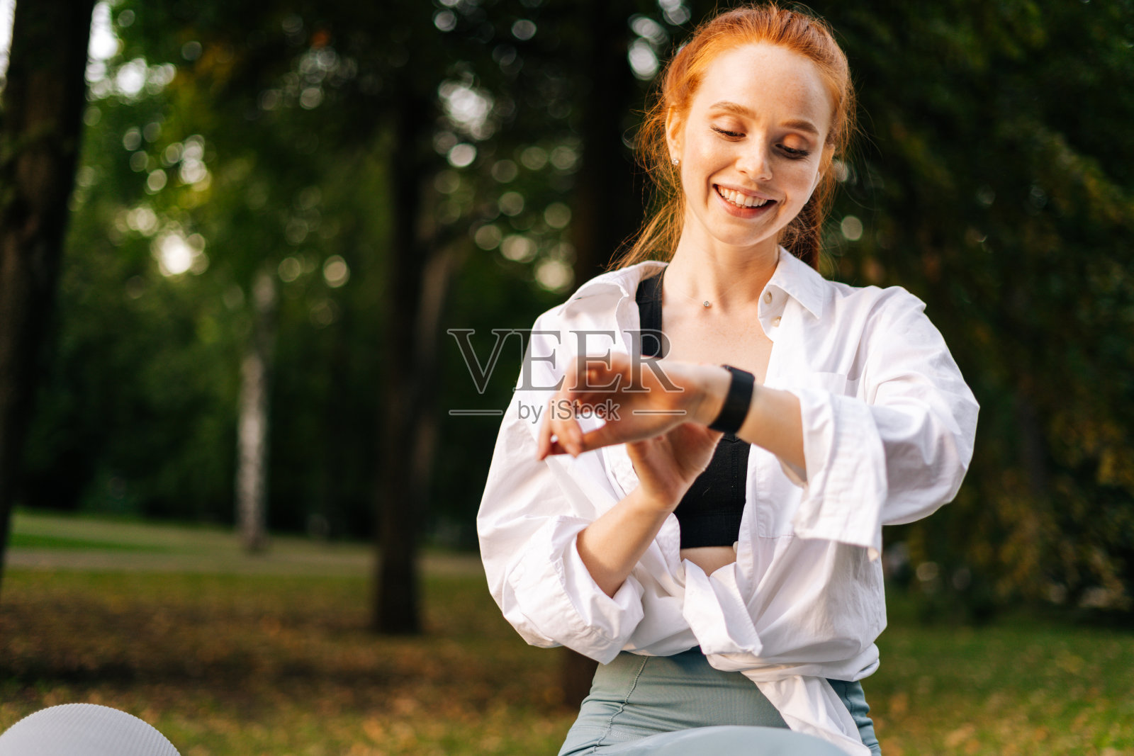 微笑漂亮的红头发的年轻女子正在使用智能手表在城市公园的户外应用程序照片摄影图片