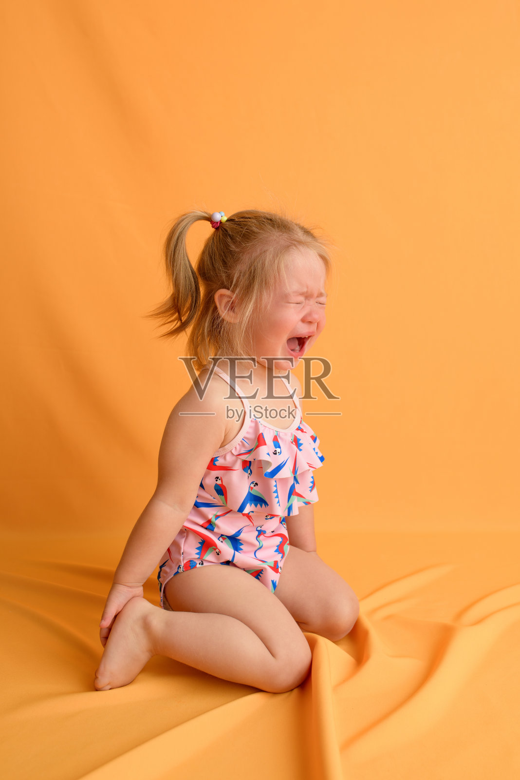 一个一岁半的小女孩坐在她的膝盖上，在黄色的背景上哭。那个女孩穿着泳衣。照片摄影图片