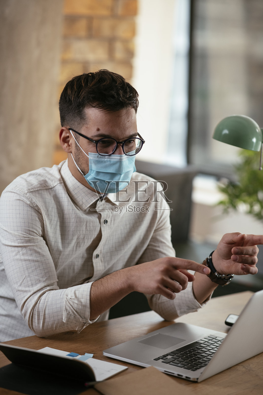 年轻的商人戴着面具在办公室里用笔记本电脑工作。照片摄影图片