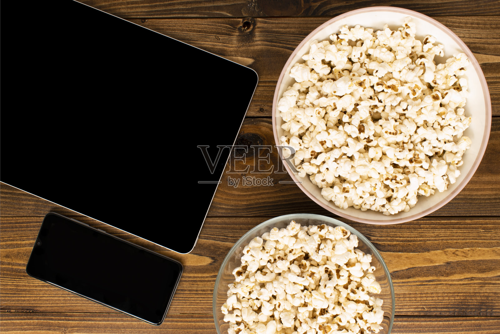 装满现代平板电脑、手机和爆米花的碗，在木地板上看电影和电视剧的时候吃。手机和平板电脑模型与黑色的拷贝空间。照片摄影图片