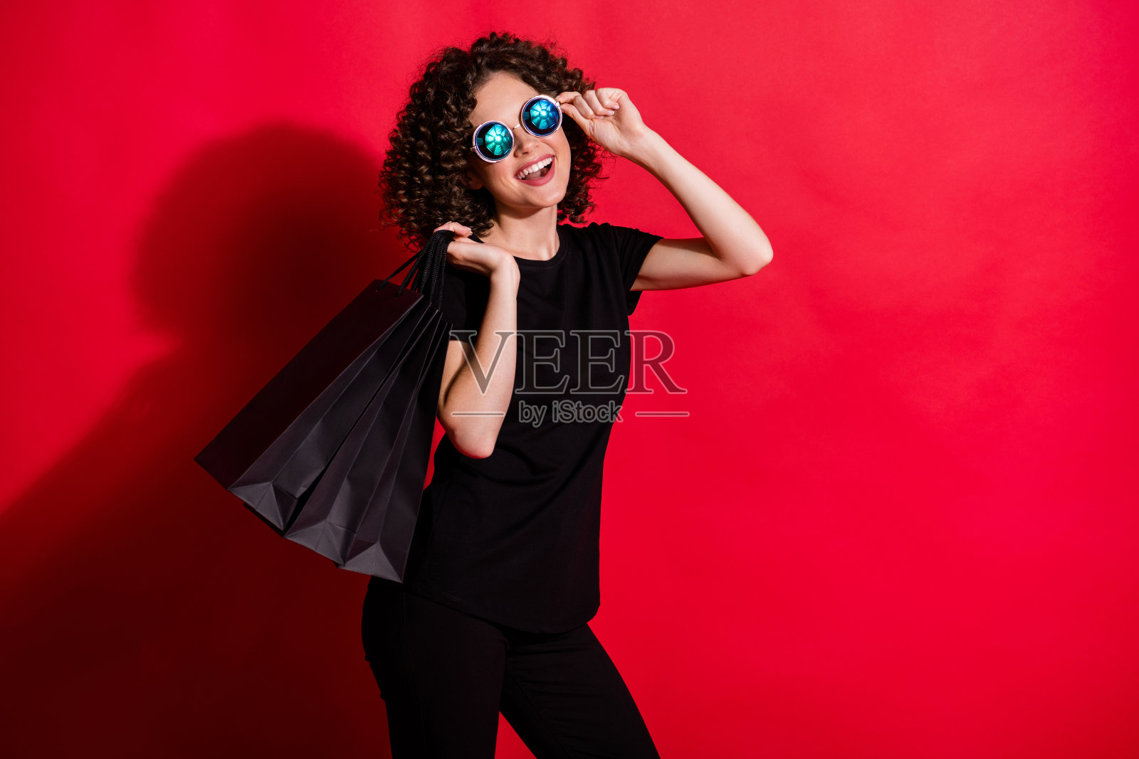 可爱的甜蜜的卷发女人的照片穿休闲的黑色装备手臂眼镜拿着包孤立的红色背景照片摄影图片