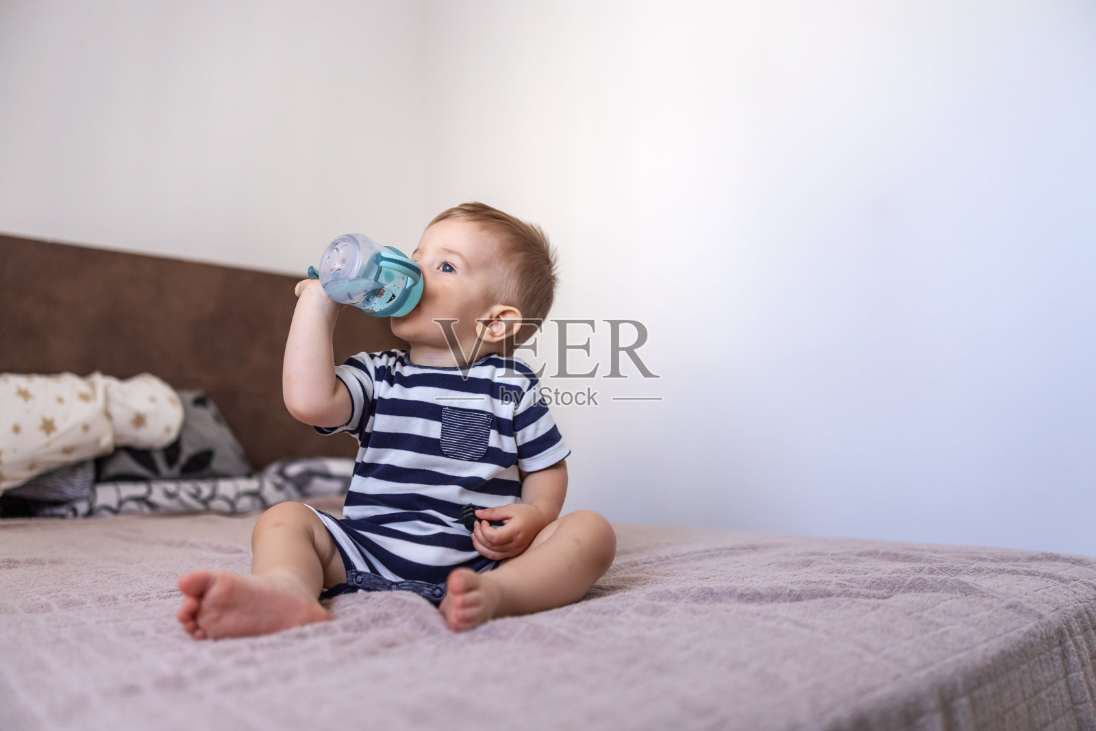 可爱的金发少年坐在卧室的床上，喝着瓶子里的水。照片摄影图片