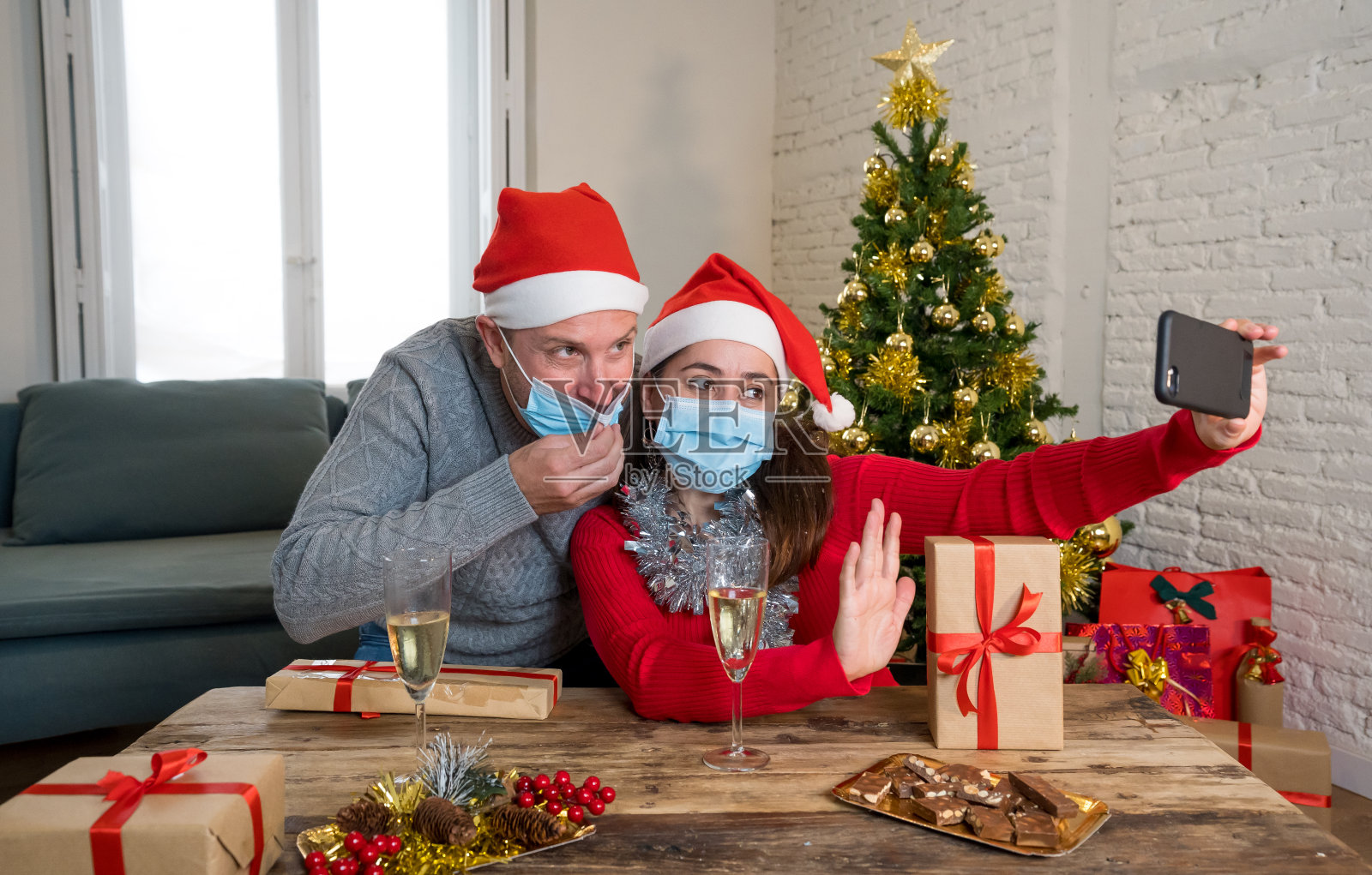 快乐的夫妇戴着面具用手机视频通话庆祝虚拟圣诞和新年派对在封锁中。社交距离、自我隔离和在线庆祝。照片摄影图片
