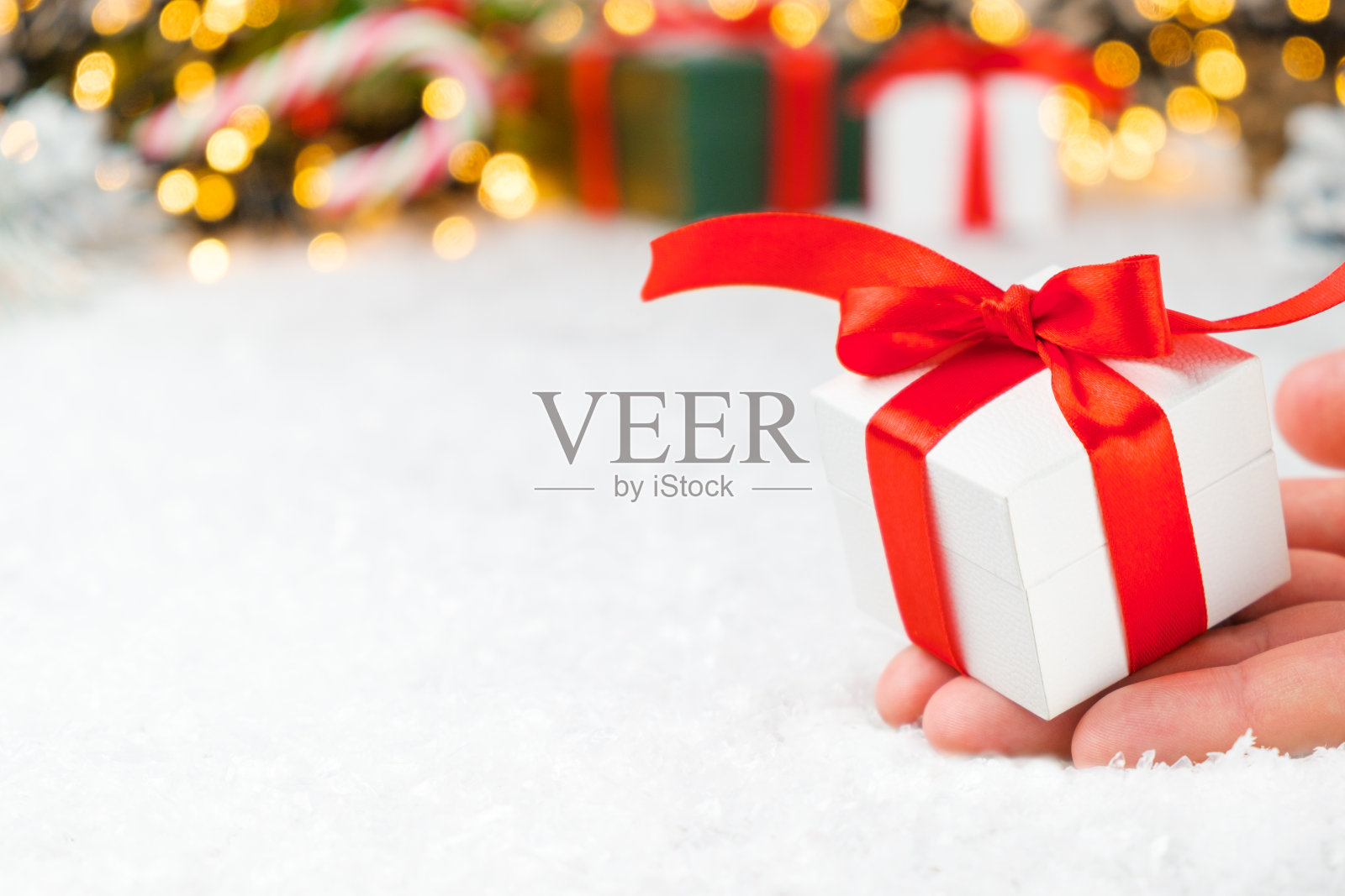 近女人手拿着一个白色的礼物盒与红色丝带的背景礼物，糖果棒，圣诞树和金色的灯与拷贝空间照片摄影图片