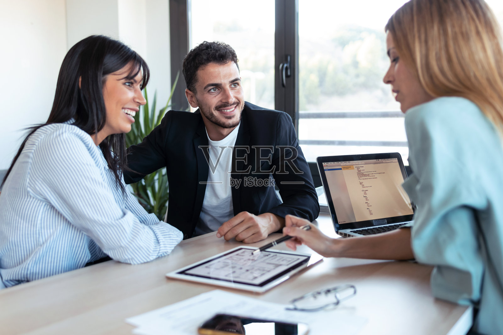 一位漂亮的房地产经纪人在办公室里一边和新婚夫妇谈买房的事，一边在平板电脑上展示房屋计划。照片摄影图片