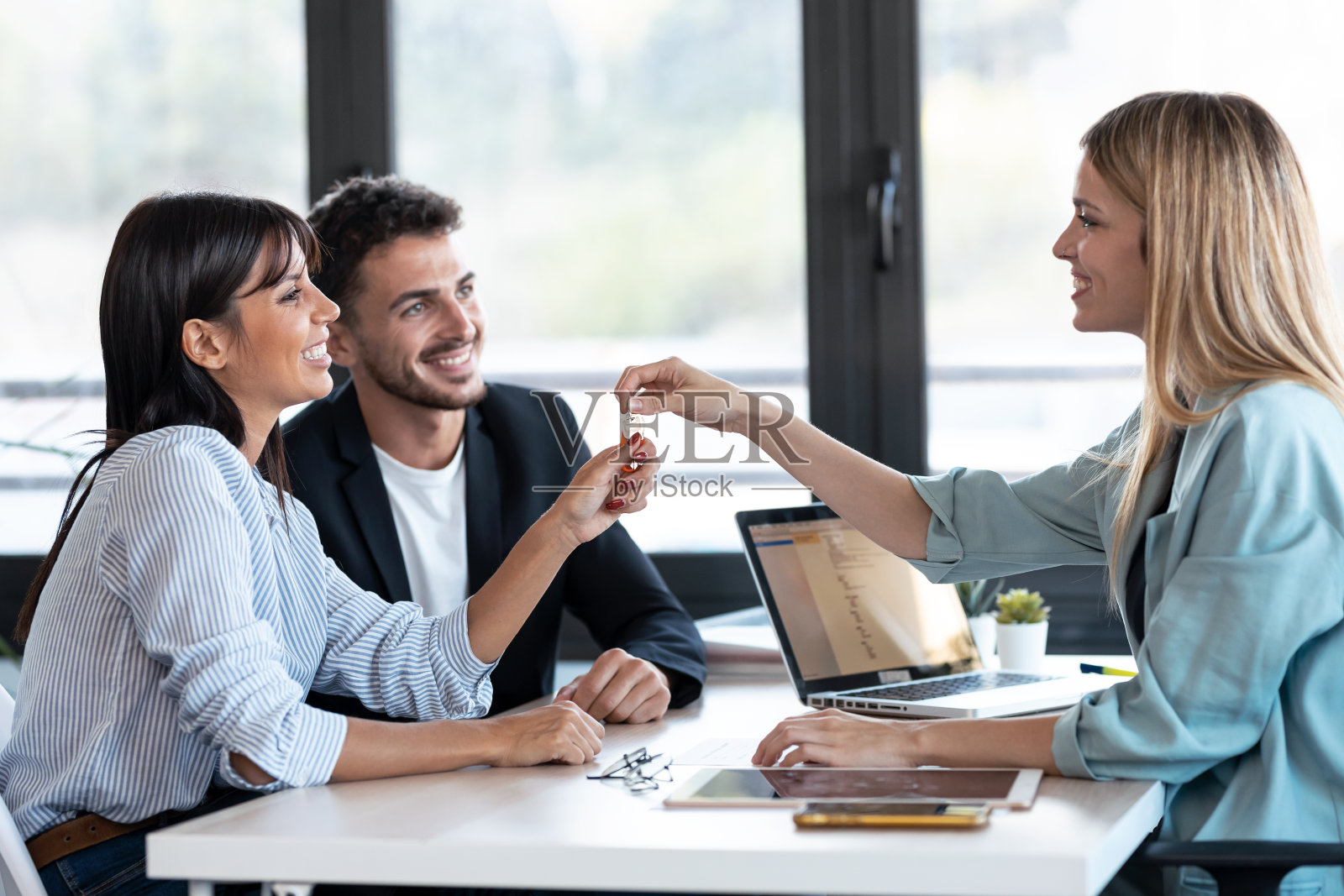 有吸引力的房地产经纪人在办公室签订合同后给年轻夫妇房子钥匙。照片摄影图片
