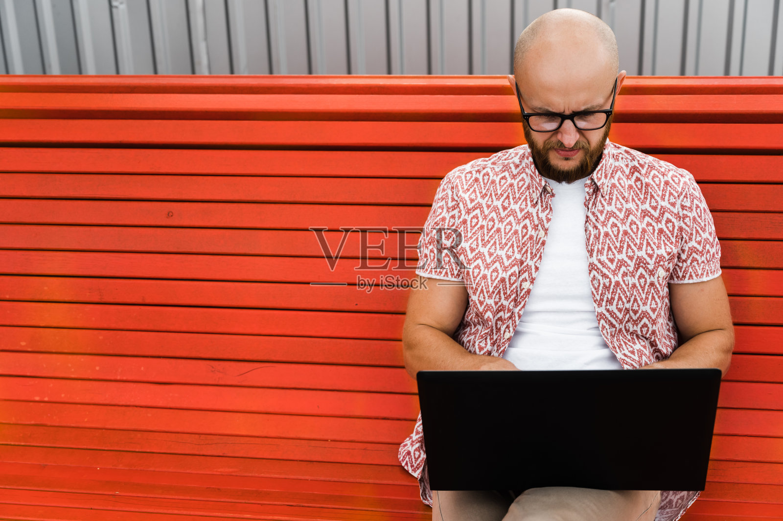 一名留着胡子、戴着眼镜、穿着时髦的白人男子坐在户外的长凳上远程使用笔记本电脑工作照片摄影图片