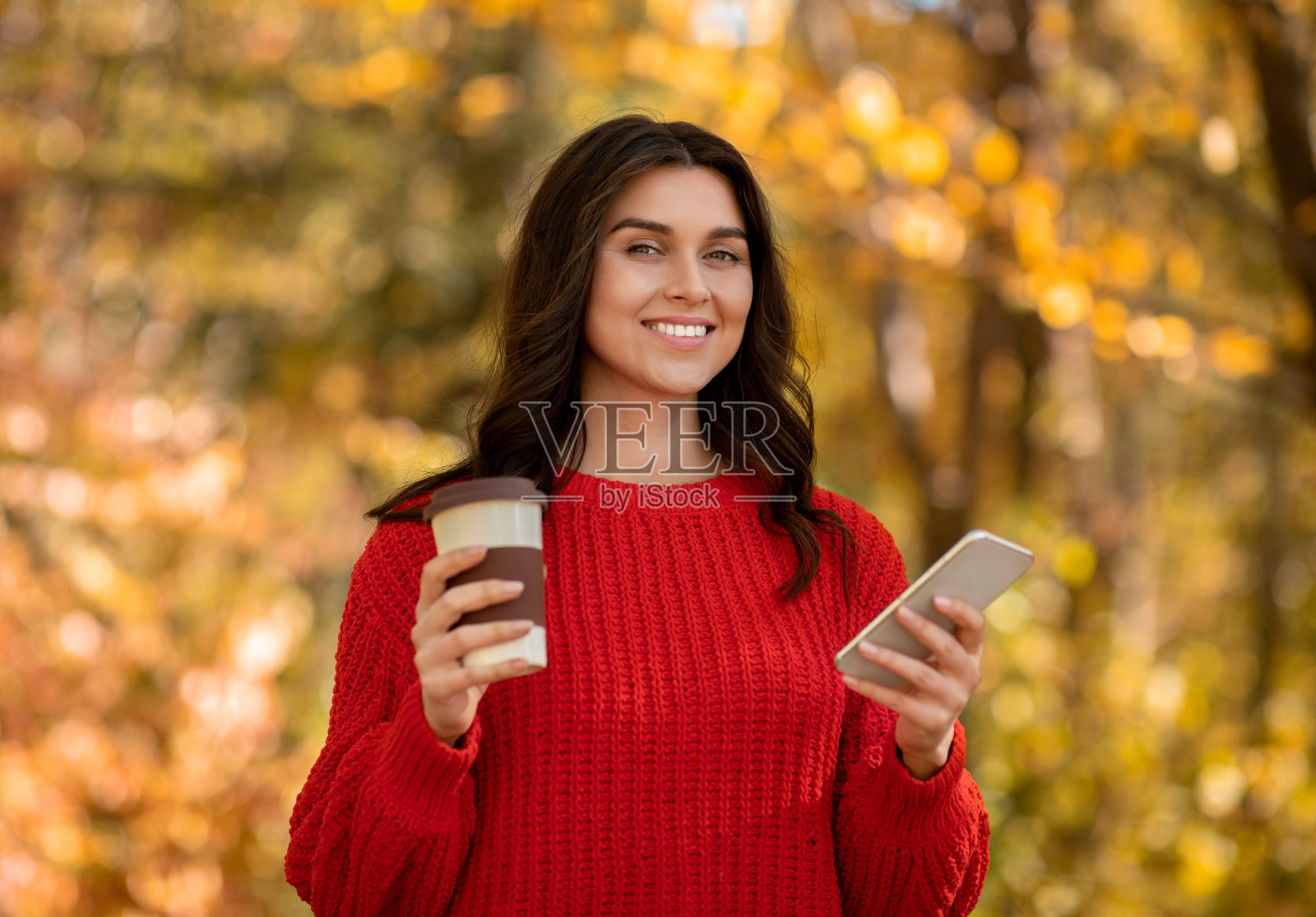 身着红色羊毛衫、手持手机、带着外卖咖啡的千禧女性在秋季公园里微笑着照片摄影图片