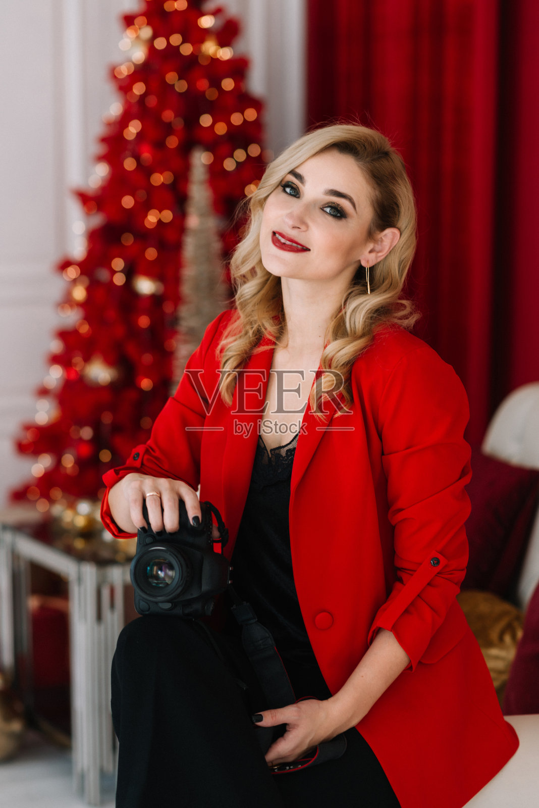 美丽的女人摄影师在红色与相机在她的手对着灯光散焦的圣诞树的背景。2021年新年快乐。软选择焦点，艺术噪音。照片摄影图片