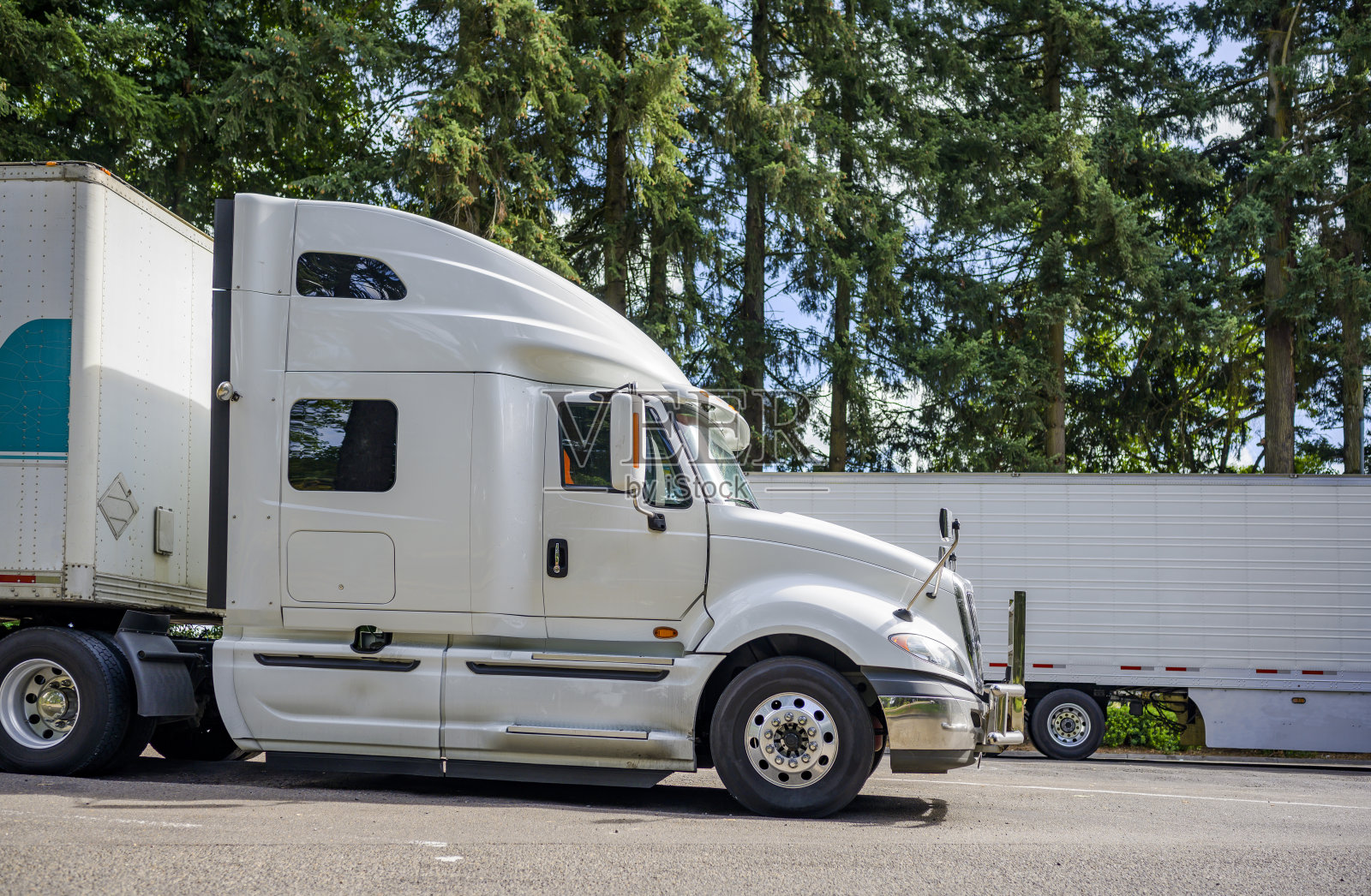 一边白色的大卡车半挂车与干货车半挂车站在休息区停车场与老森林树照片摄影图片