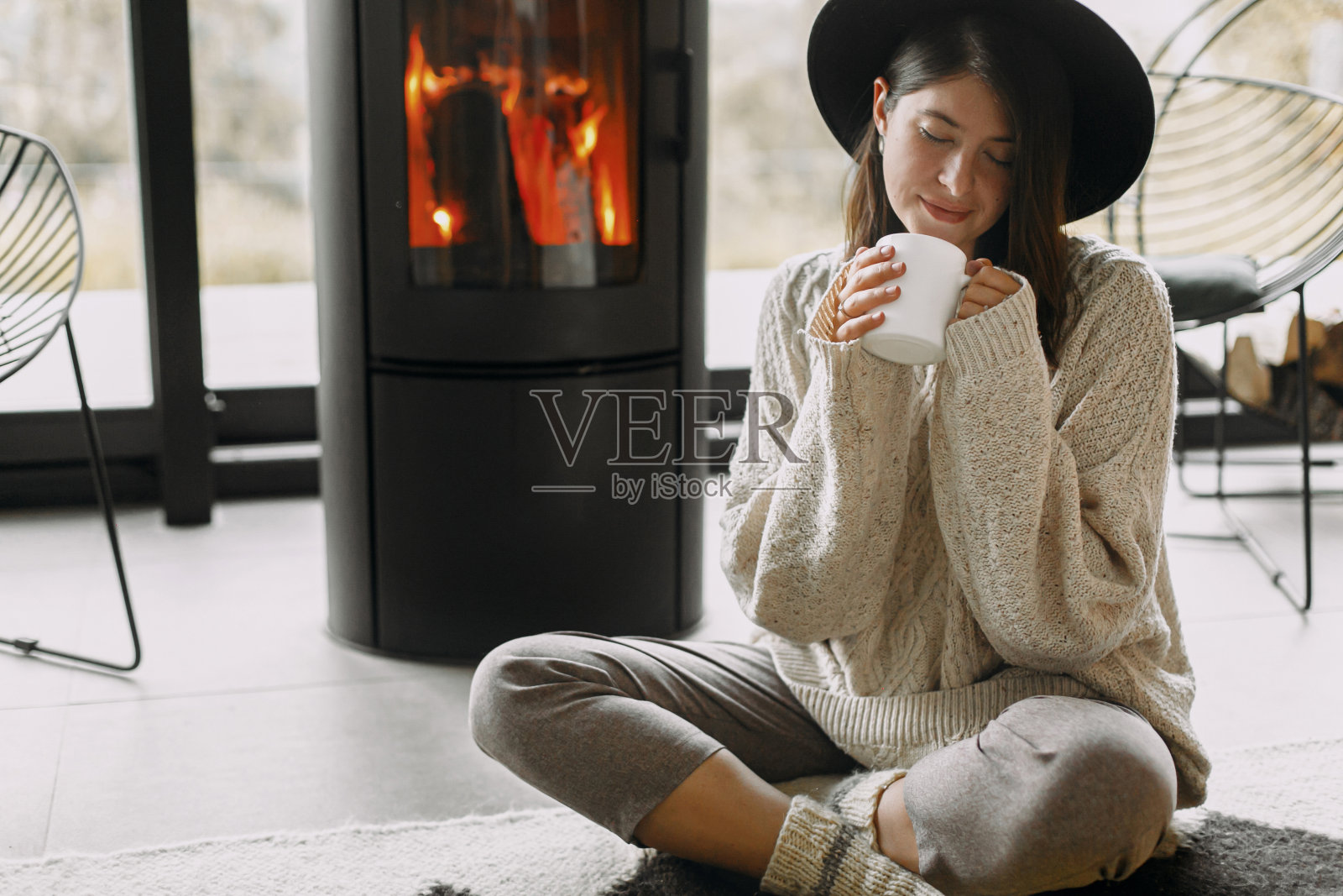 时尚的女人在针织毛衣和帽子拿着温暖的茶和放松在现代壁炉照片摄影图片