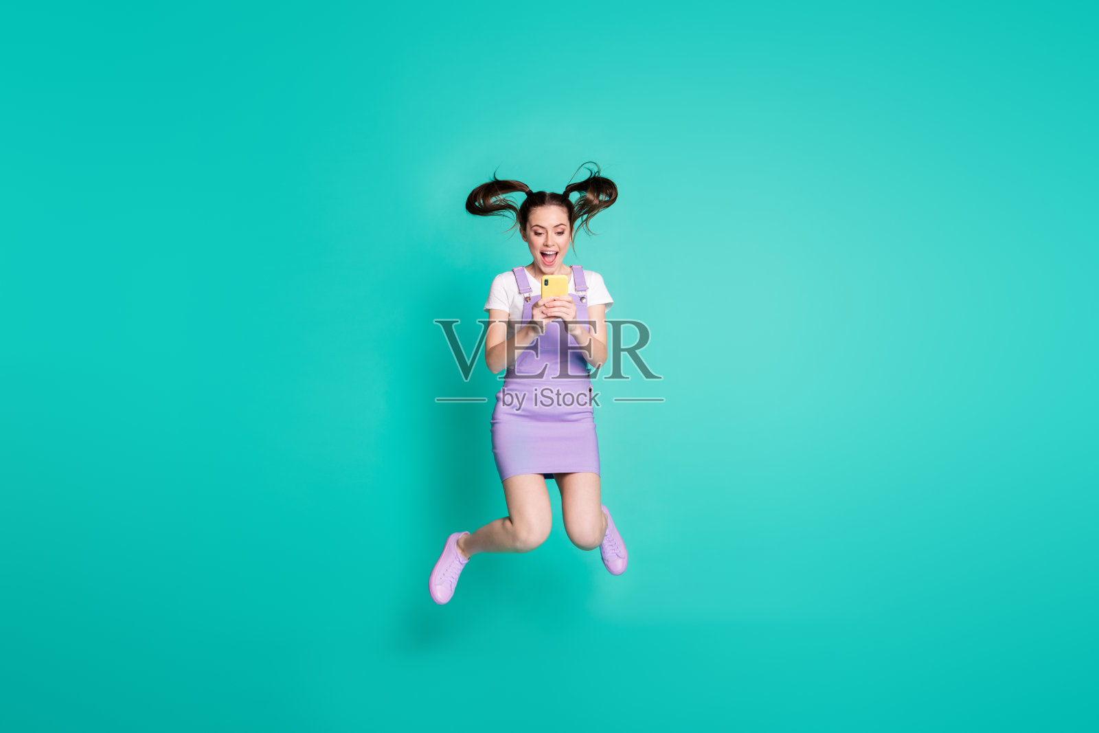 全身大小的照片，黑发女孩的尾巴跳高使用智能手机社交媒体孤立在生动的蓝绿色背景照片摄影图片