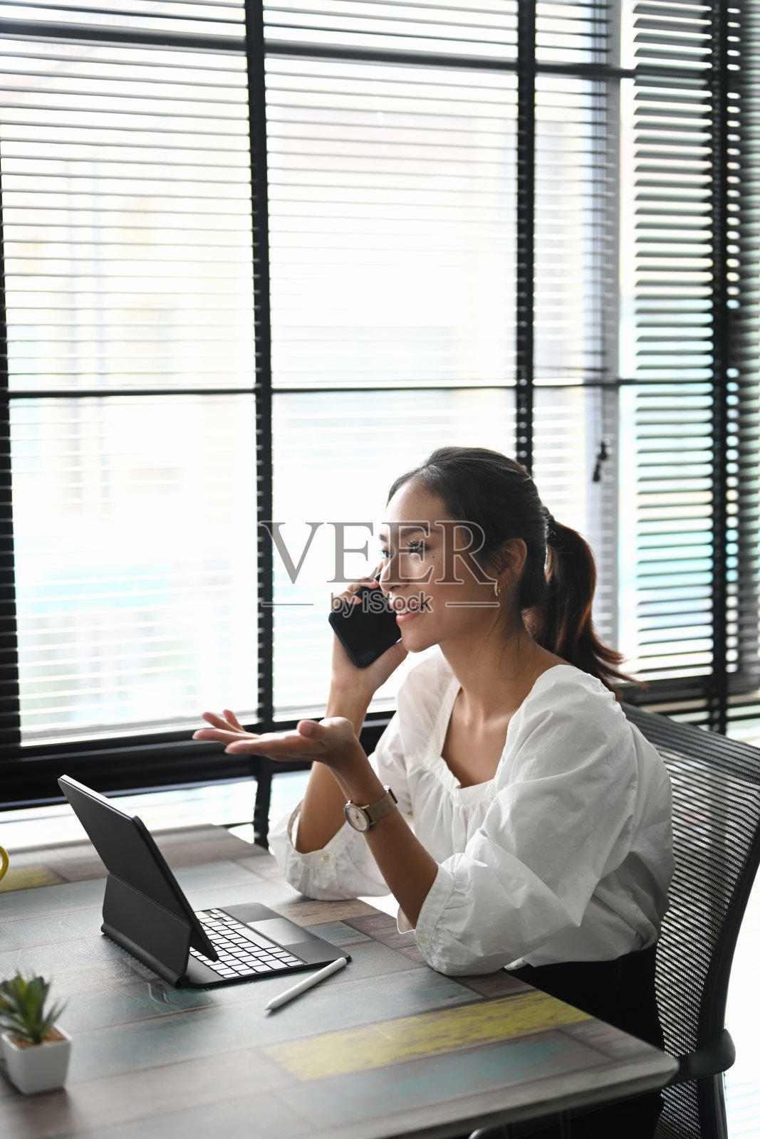 一个愉快的年轻女商人正坐在她的工作场所用手机和平板电脑聊天。照片摄影图片