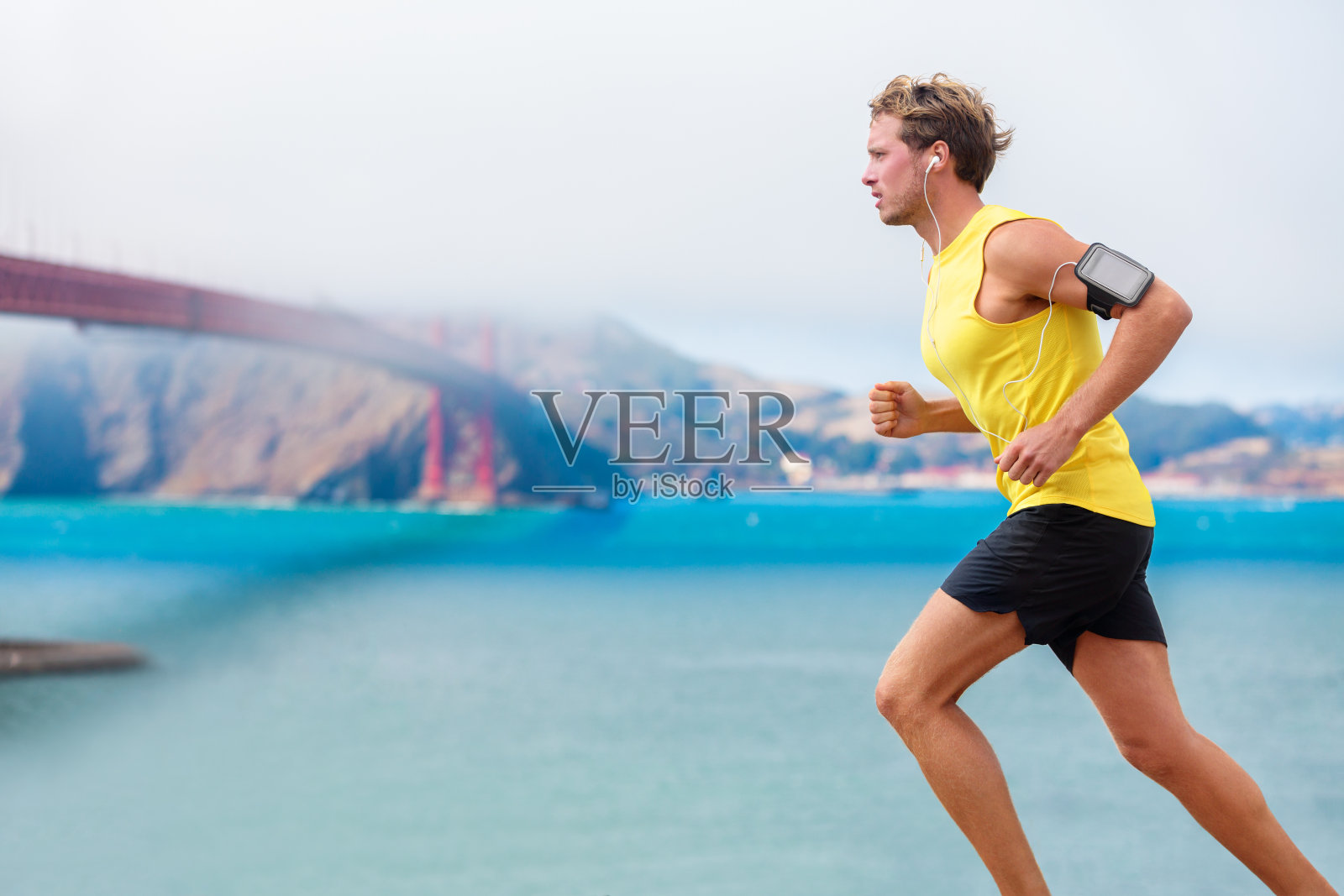 跑步者在跑步时听音乐。跑步运动员在智能手环和耳机上使用应用程序。运动健康的年轻慢跑者在旧金山湾和金门大桥慢跑。照片摄影图片