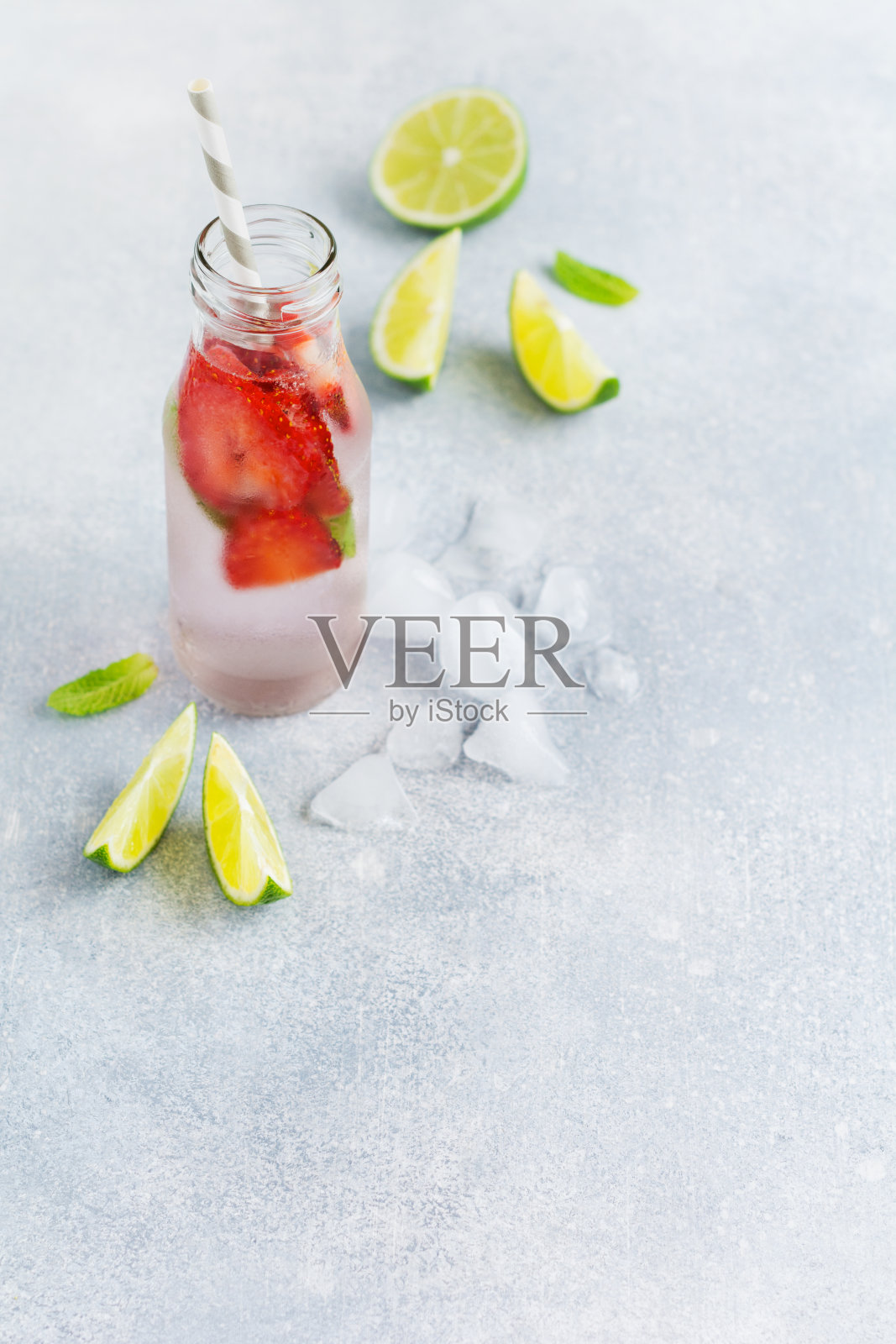 夏天的柠檬水，冰，草莓，薄荷和酸橙装在一个瓶子和一个纸管在混凝土灰色的背景。健康饮酒的概念。照片摄影图片