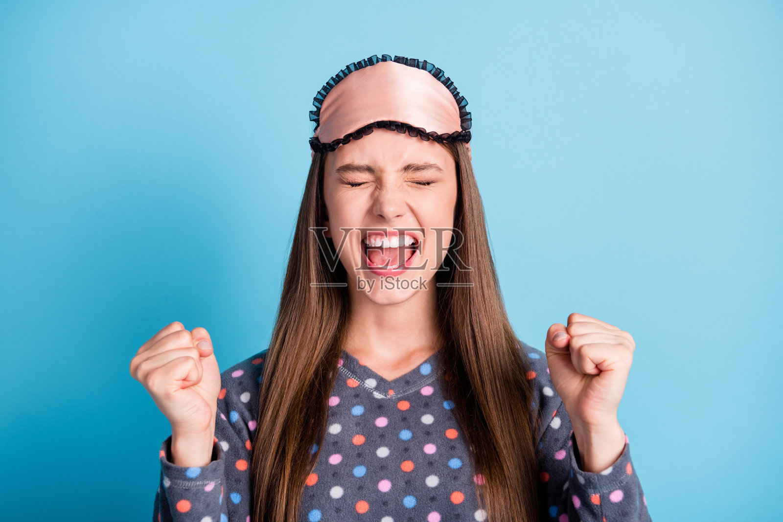 特写大头照片的可爱的模特年轻的女孩举起拳头，张开嘴，闭上眼睛中奖穿面具点衬衫睡衣睡衣孤立的蓝色背景照片摄影图片
