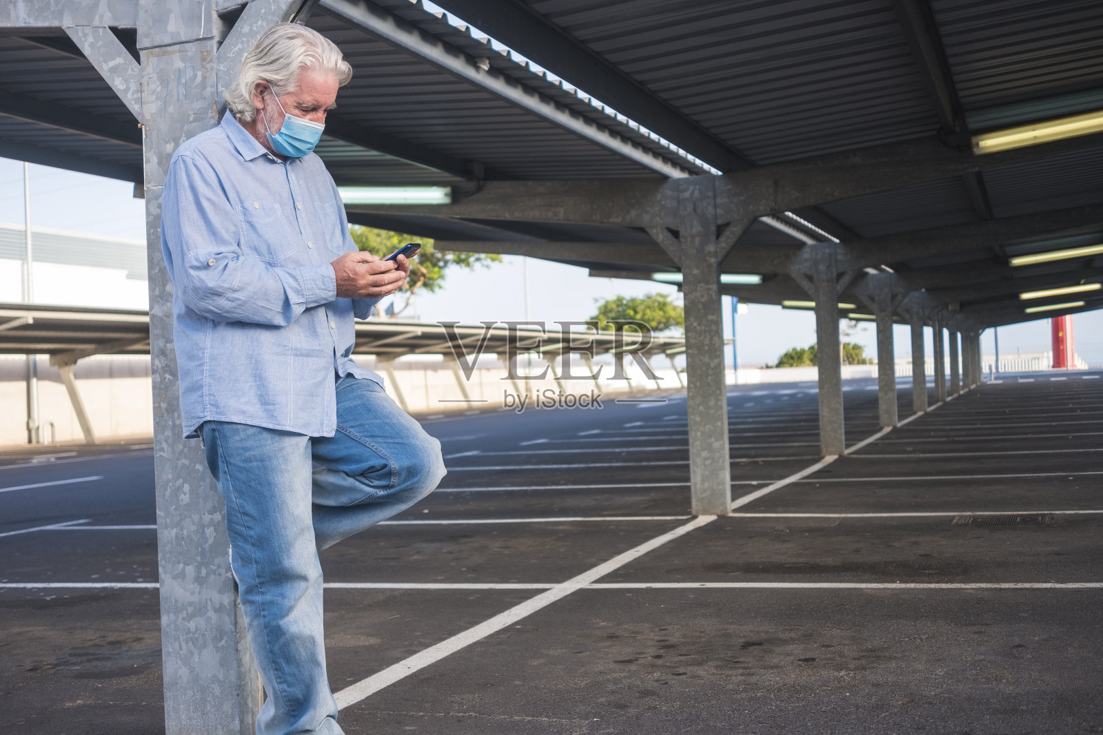 冠状病毒一名因冠状病毒戴着医用口罩的老人站在一个废弃停车场的金属结构下看他的智能手机。没有其他人照片摄影图片