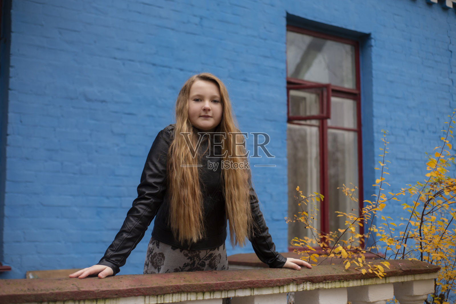 一个穿着皮夹克和黑色帽子的可爱的黑发少女的肖像，背景是青砖房子和旧篱笆。舒适、时尚、风格照片摄影图片