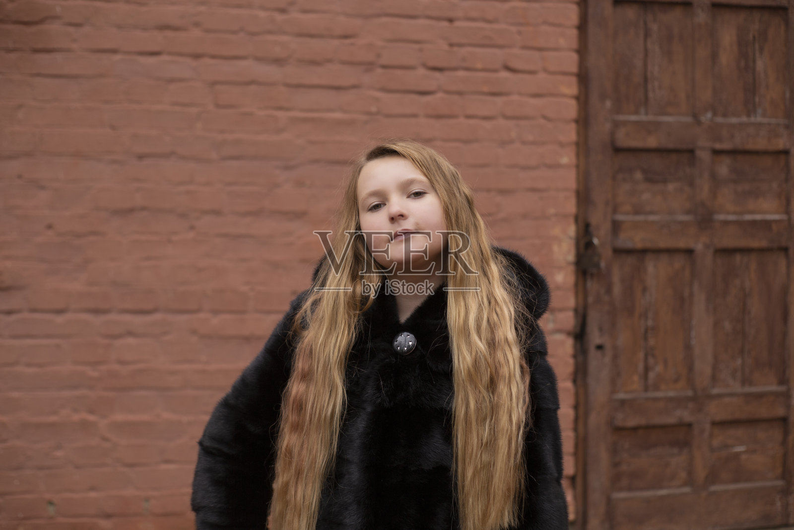 可爱的黑发少女的肖像在棕色的砖墙和旧木门的背景黑色毛皮大衣。舒适、时尚、风格照片摄影图片