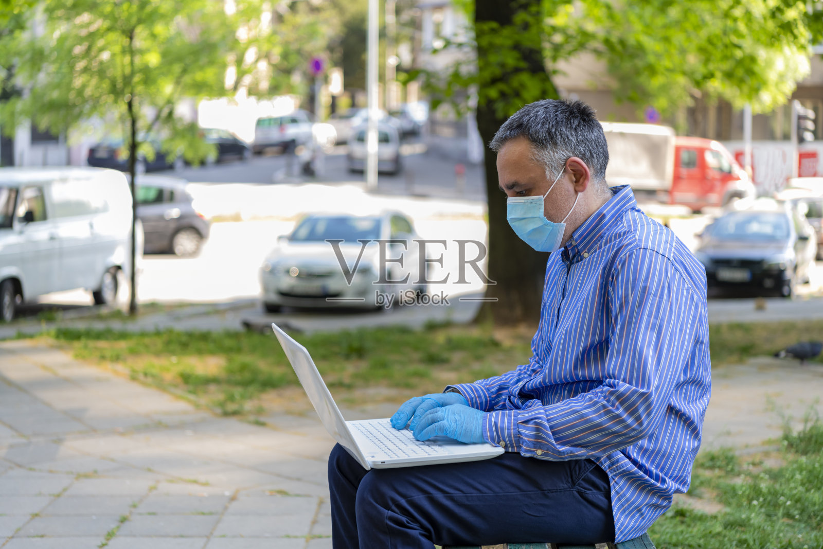 穿着衬衫的英俊商人戴着防护面具和手套在公园里操作他的笔记本电脑。照片摄影图片