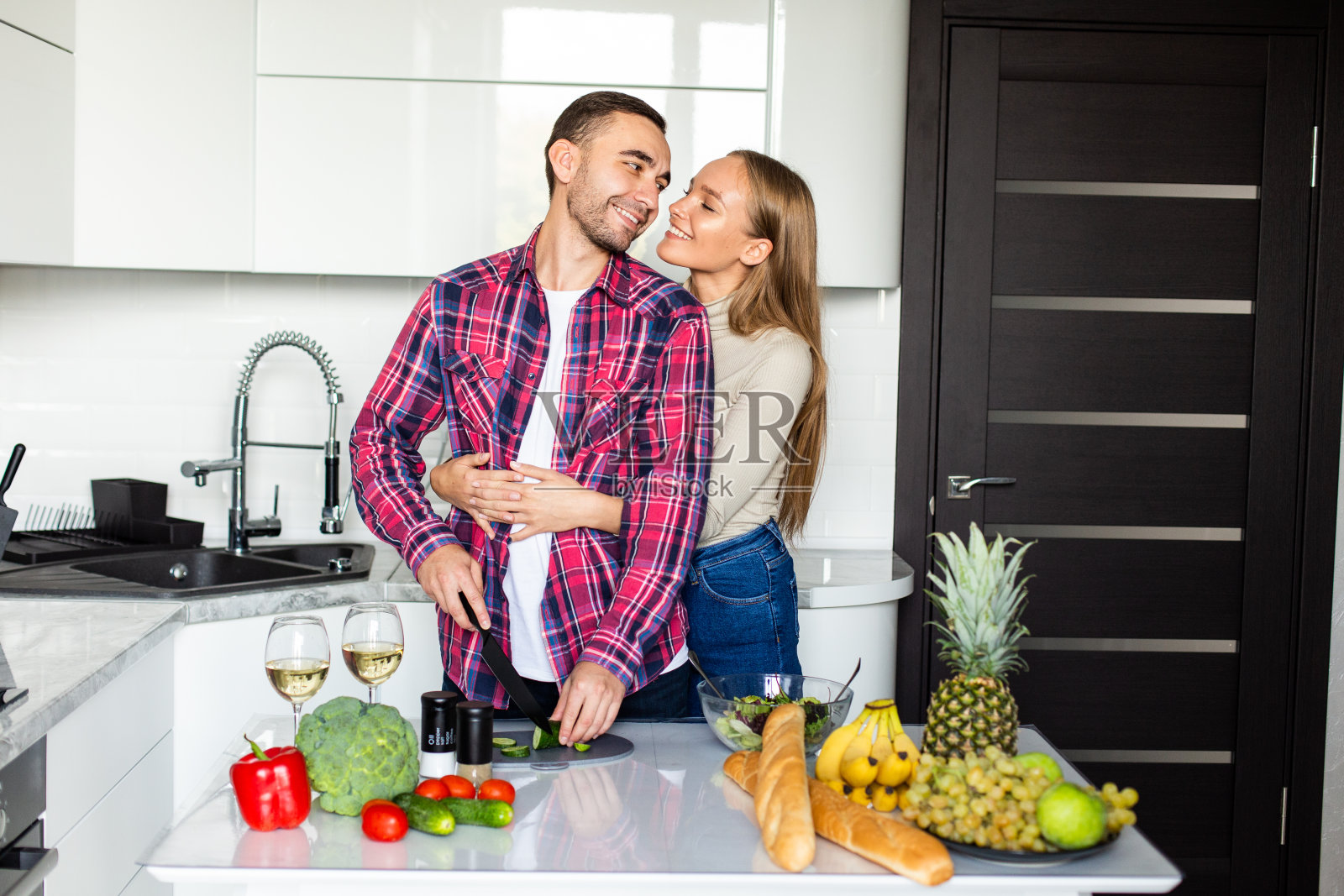 一名年轻男子亲吻他的妻子，妻子正在厨房里搅拌沙拉配料照片摄影图片