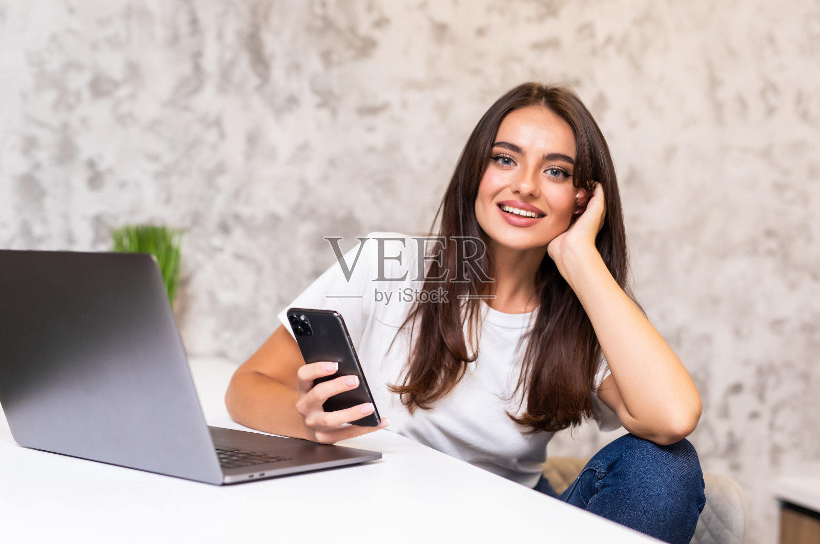 年轻女子使用电话和笔记本电脑，自由职业者在家在线工作。自信的年轻女性看着智能手机屏幕，阅读信息或电子邮件照片摄影图片