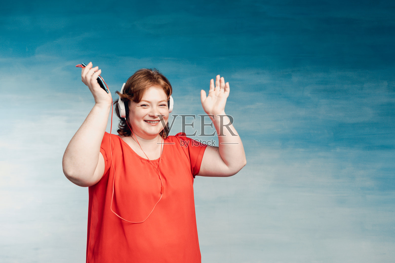 年轻迷人的红发胖女人戴着赏心悦目的耳机在蓝色背景下听音乐。戴着耳机愉快地听着音乐，微笑着照片摄影图片