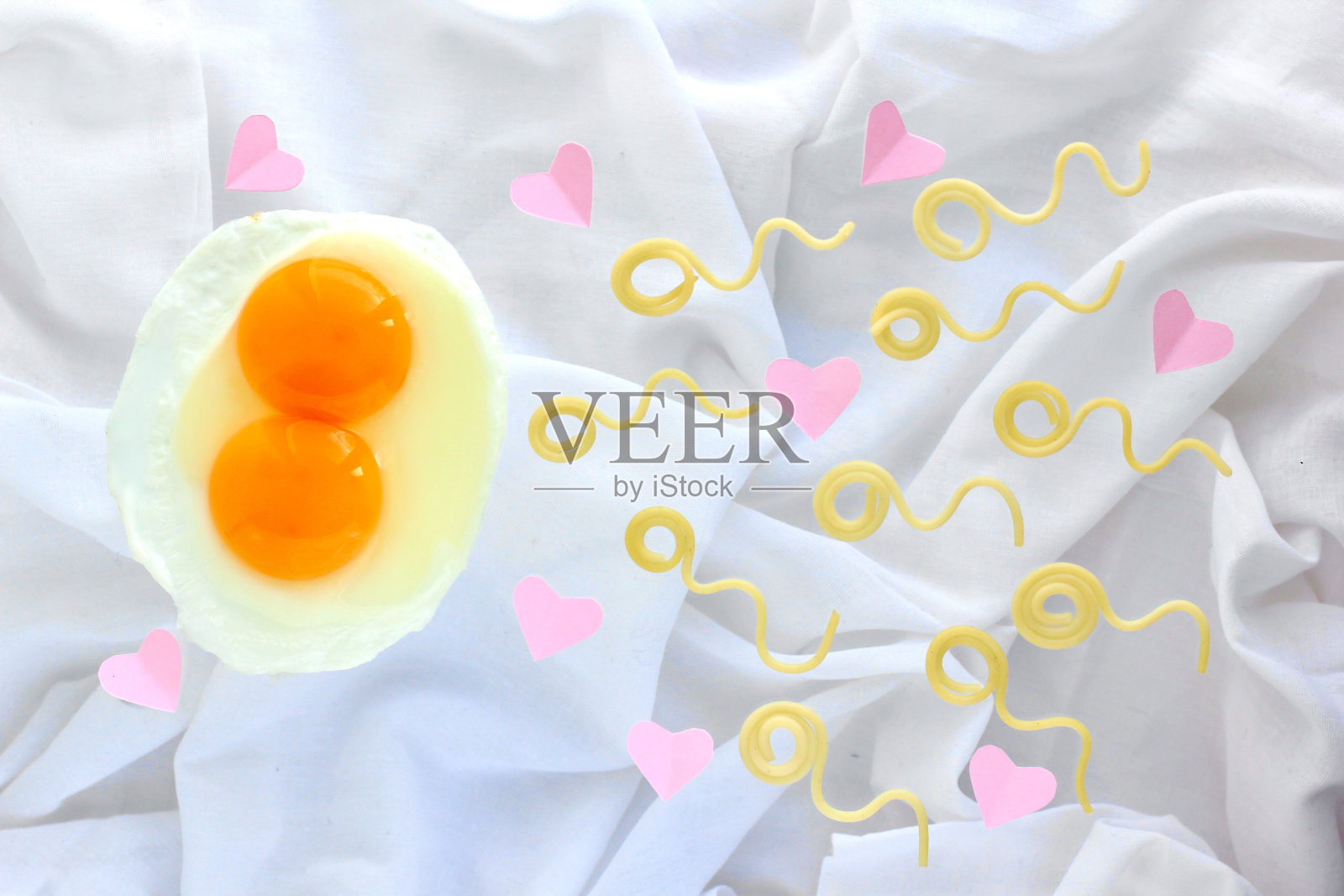 两个蛋黄的鸡蛋和意大利面看起来像精子竞争，精子悬浮在白色床单和粉红色的心的背景上。时髦的概念，受精，双胞胎，爱情，情人节。照片摄影图片