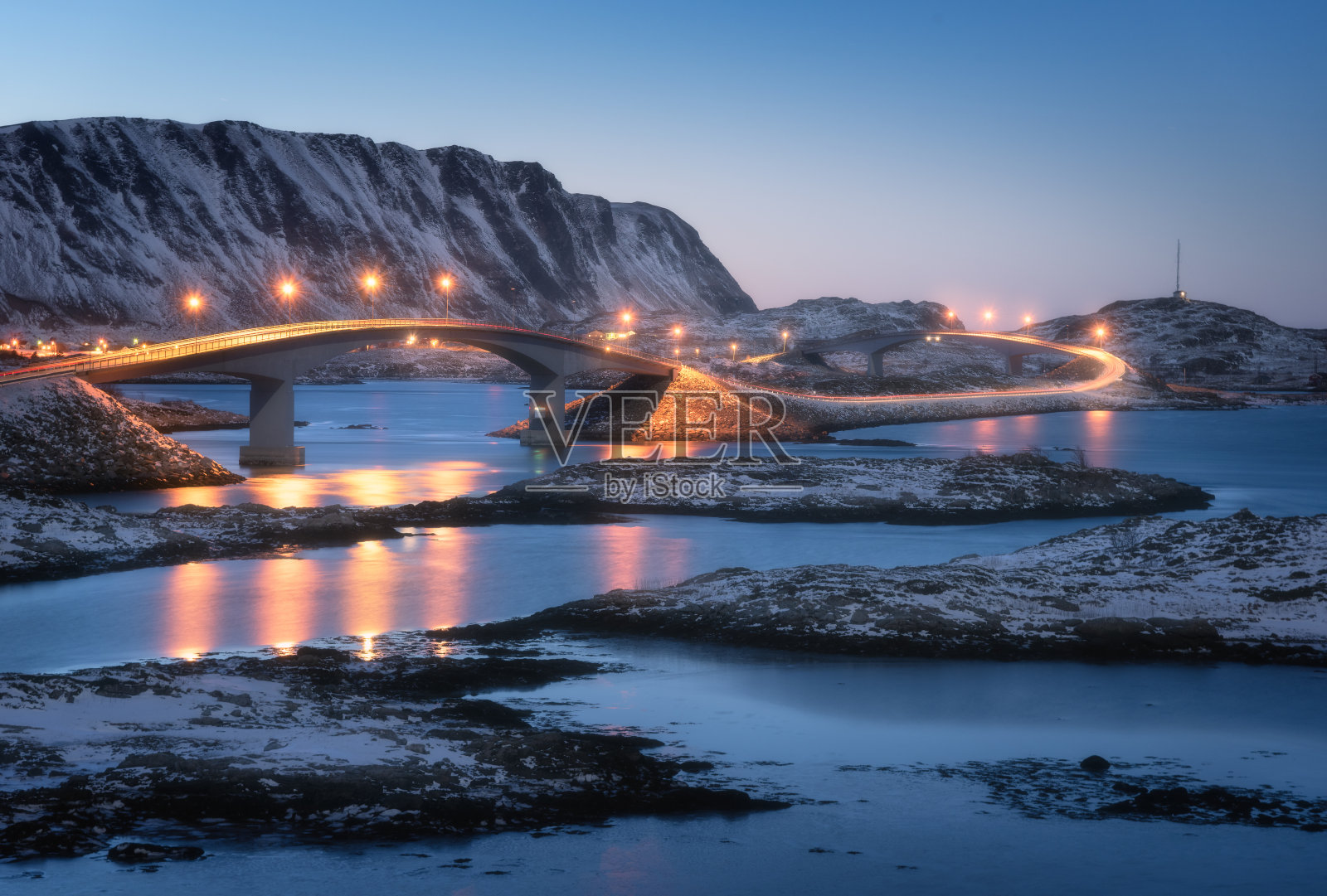 桥上灯火通明，雪山覆盖，村庄和蓝天映在水中。夜景有桥、雪石、城市灯光、大海。挪威罗浮敦群岛的冬天。路照片摄影图片