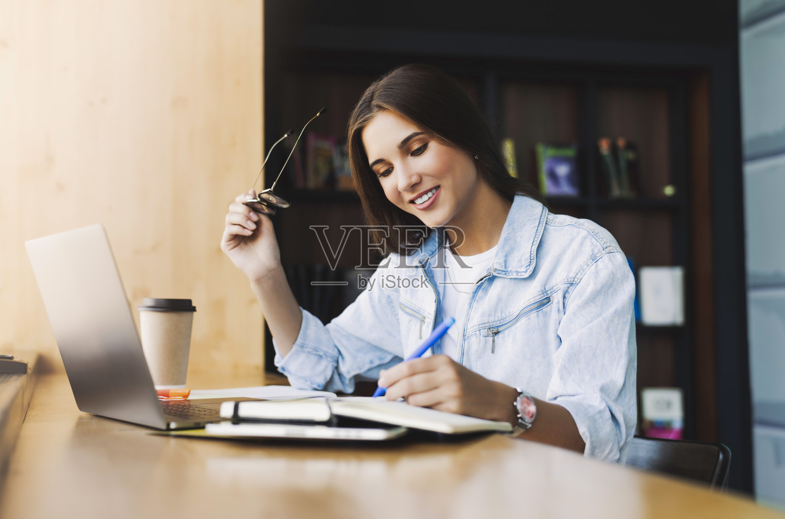 有魅力的女商人正坐在桌子前的笔记本电脑前。漂亮的黑发女孩微笑着，拿着眼镜照片摄影图片