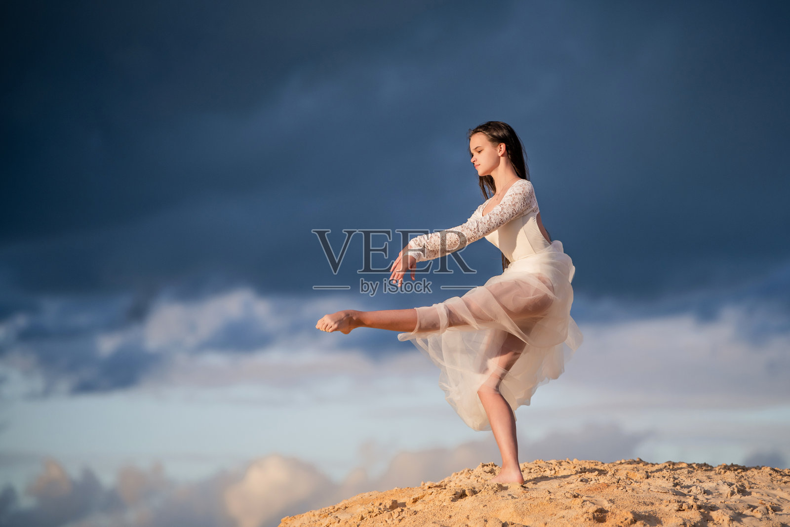 年轻的芭蕾舞女演员穿着一件白色的长裙，像一只鸟一样站在阿拉贝斯克，她的裙子在暴风雨的夜空的背景下展开。照片摄影图片