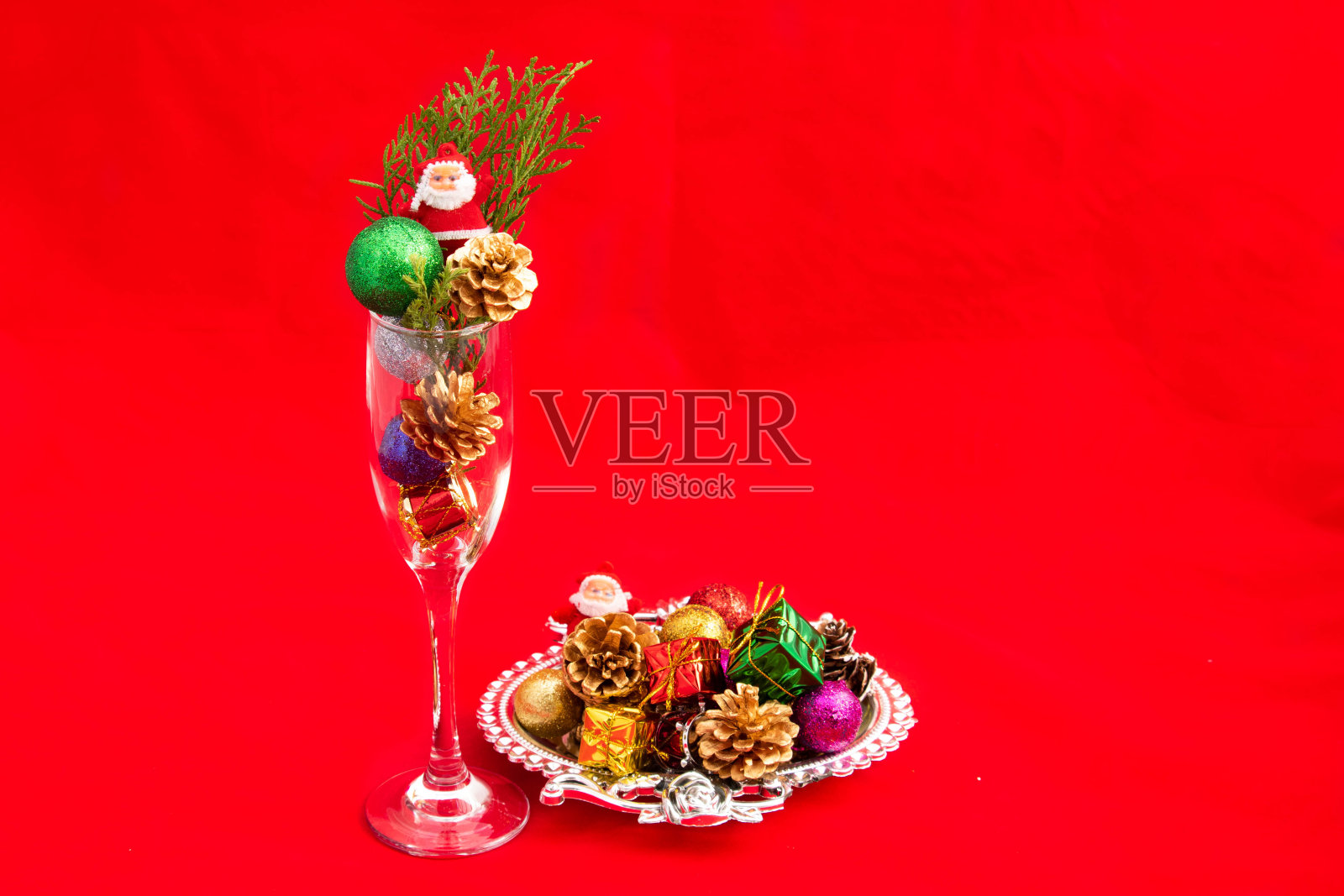 装饰装饰松果，闪光球，小圣诞老人礼盒安排在酒杯和银色设计师托盘与浪漫的圣诞主题明亮的红色背景照片摄影图片