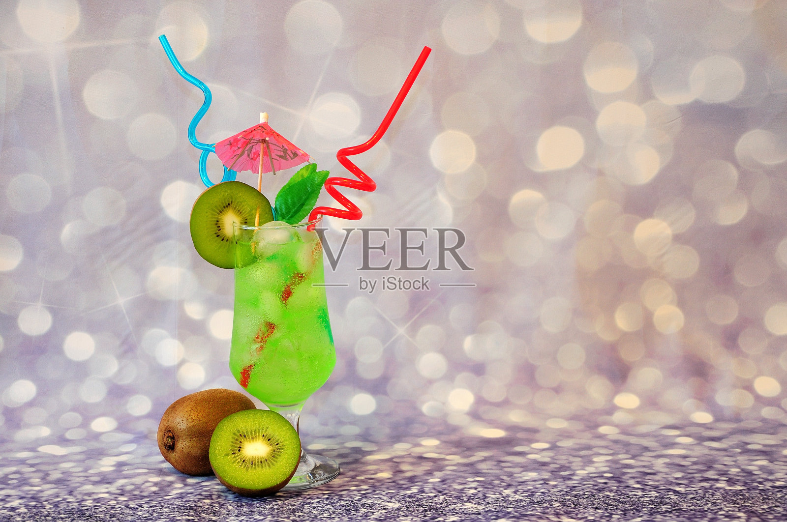 一杯加冰的绿果汁，吸管，鸡尾酒伞和成熟的猕猴桃在灰色光泽的背景。照片摄影图片