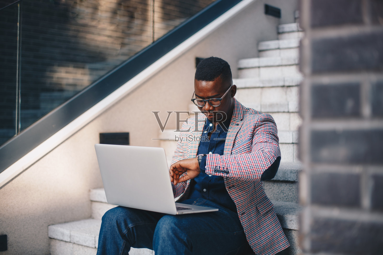 一个年轻的非洲裔美国人坐在楼梯上，把他的手提电脑放在他的膝盖上，看着手表。照片摄影图片