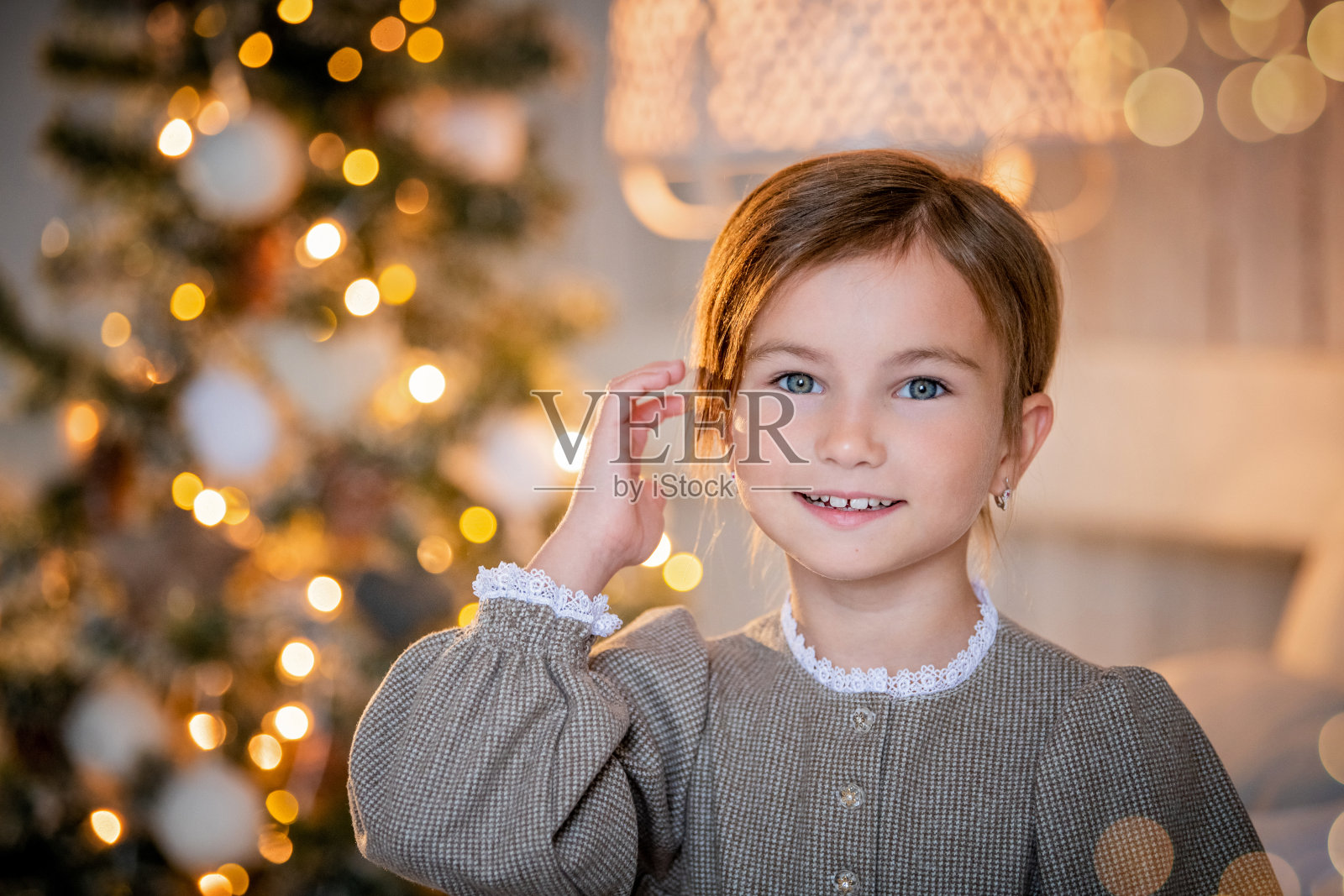 一个小女孩的肖像，微笑着从圣诞树后面窥视。这棵树矗立在家里一个舒适的房间里，被花环装点得熠熠生辉照片摄影图片
