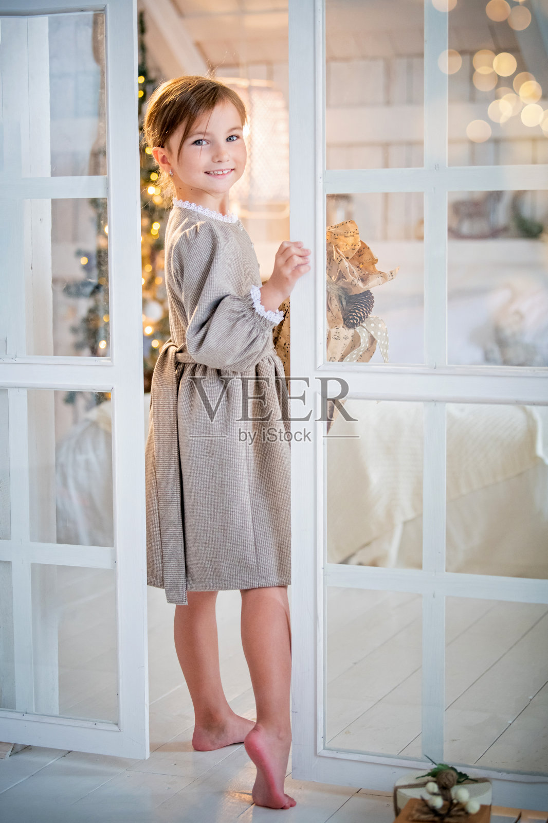 在一个明亮的室内女孩高兴地看着门外，笑了。透过玻璃可以看到一棵圣诞树。照片摄影图片