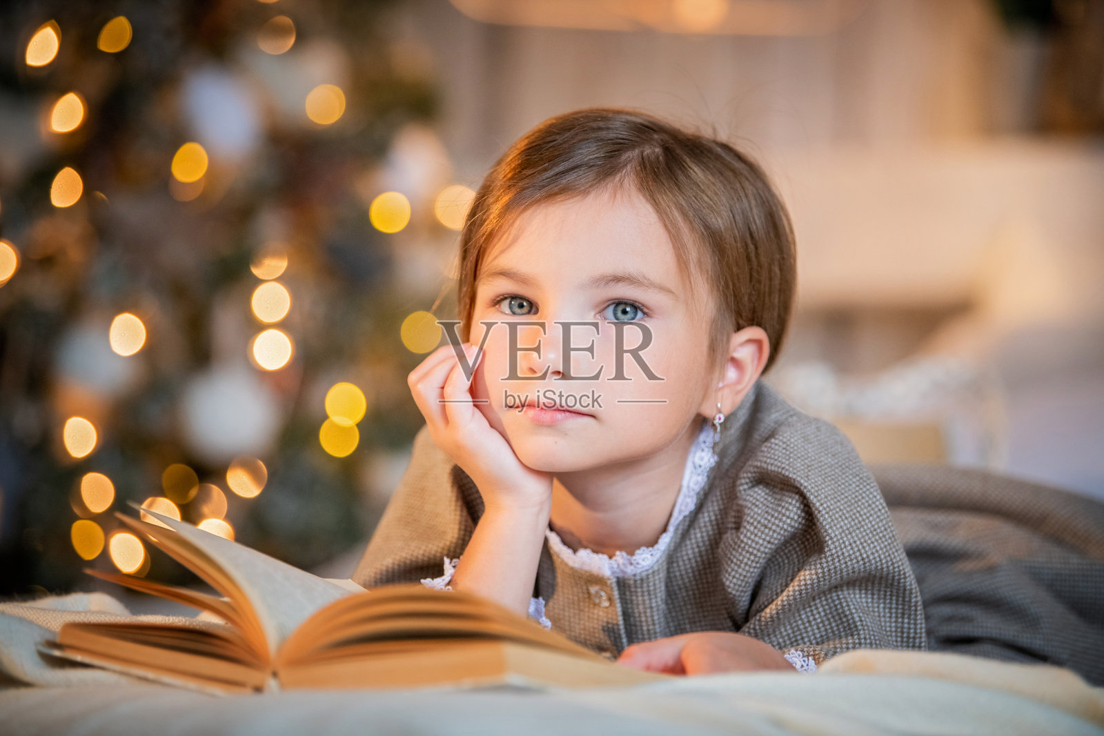 小女孩躺在明亮的沙发上看书，背景是一棵闪烁的圣诞树照片摄影图片