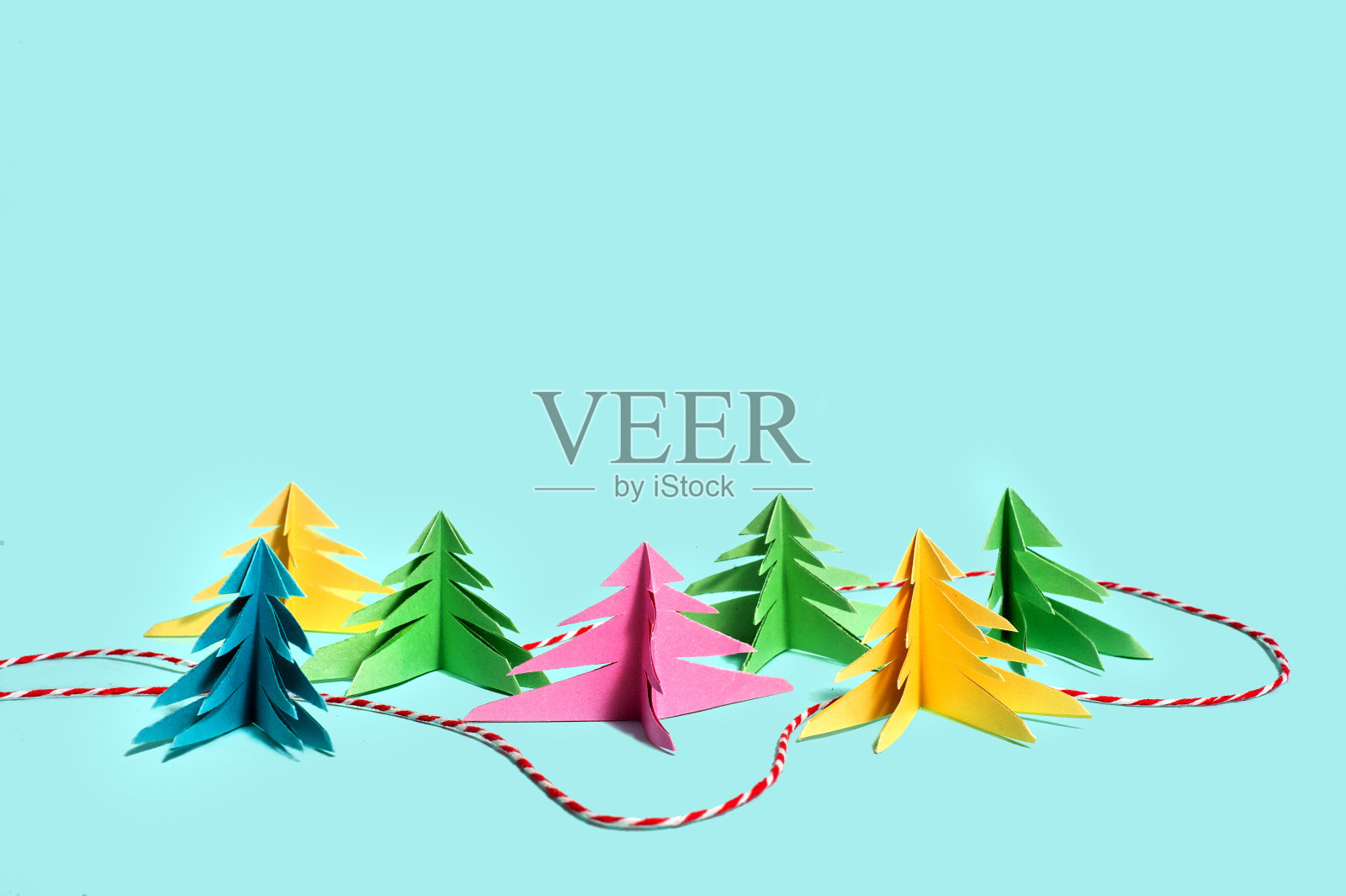 五颜六色的纸树映衬着蓝色的背景，在它们之间，就像森林里的一条小路，铺着一条红白相间的圣诞线。照片摄影图片